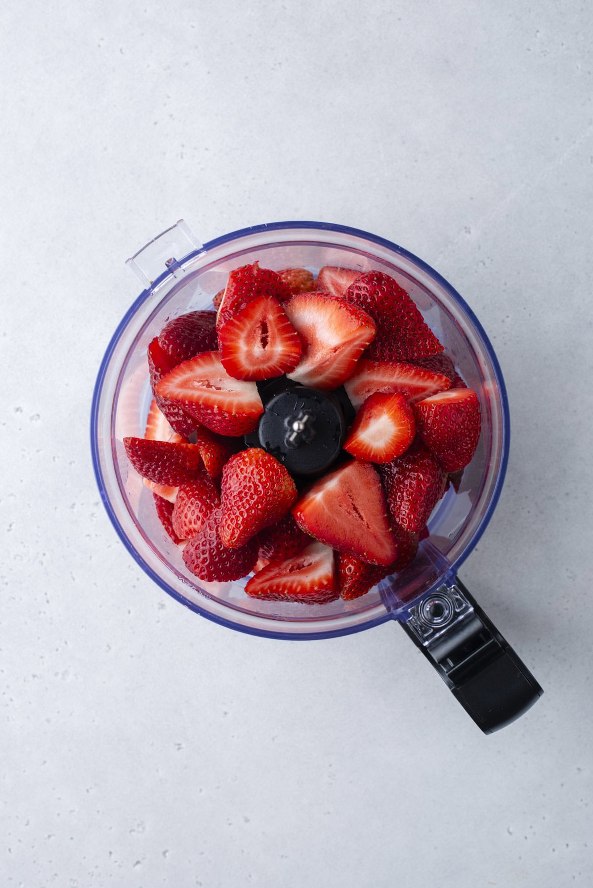 Top view of a food processor filled with fresh, halved strawberries on a light gray surface.