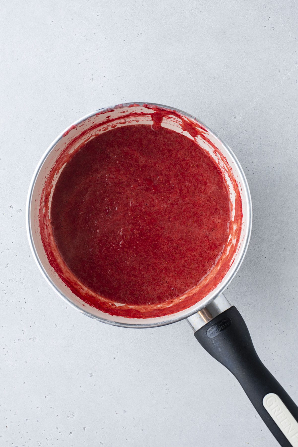 A saucepan with a red, smooth fruit sauce or puree inside, resting on a light gray surface. The sauce coats the sides of the pan, and the handle is black with a brand name visible.