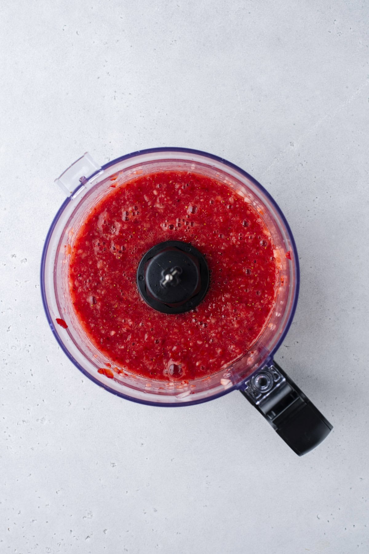 A food processor filled with a bright red, blended mixture, likely fruit puree or sauce, sits on a light gray countertop.