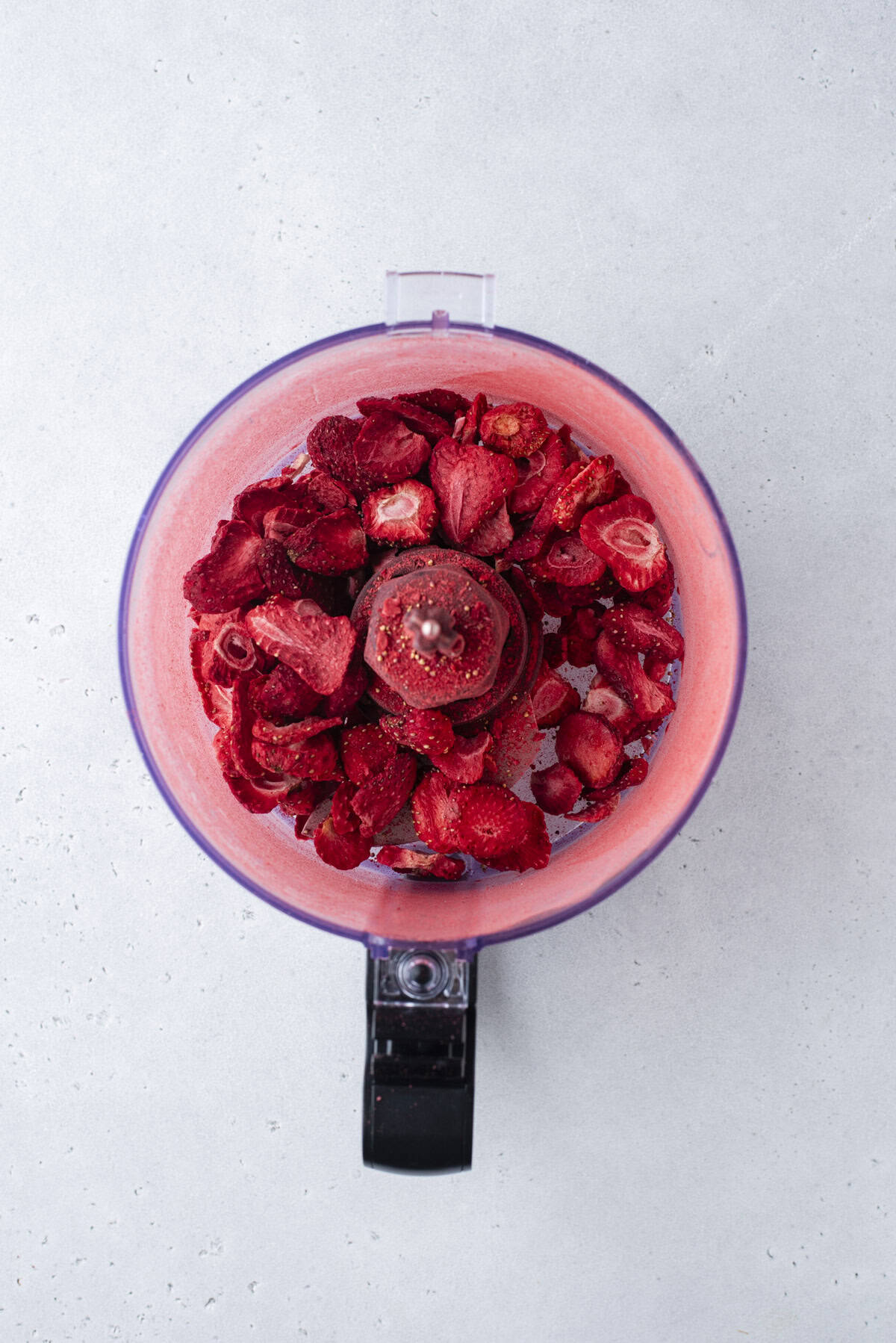 A food processor bowl filled with freeze-dried strawberries, viewed from above on a light gray surface.