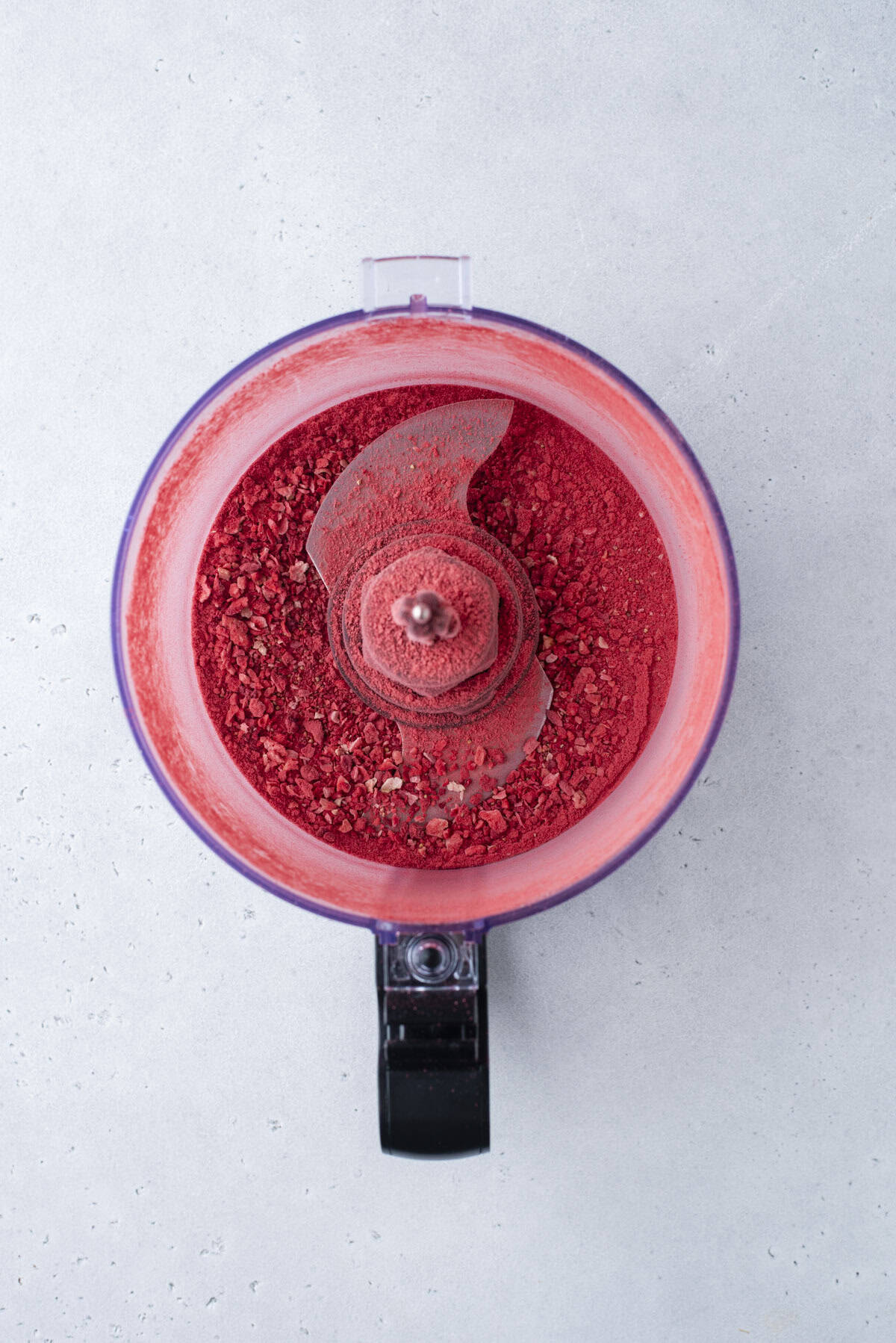A food processor with bright red powder inside sits on a light gray surface, viewed from above. The lid is off, revealing the metal blade and finely ground contents.