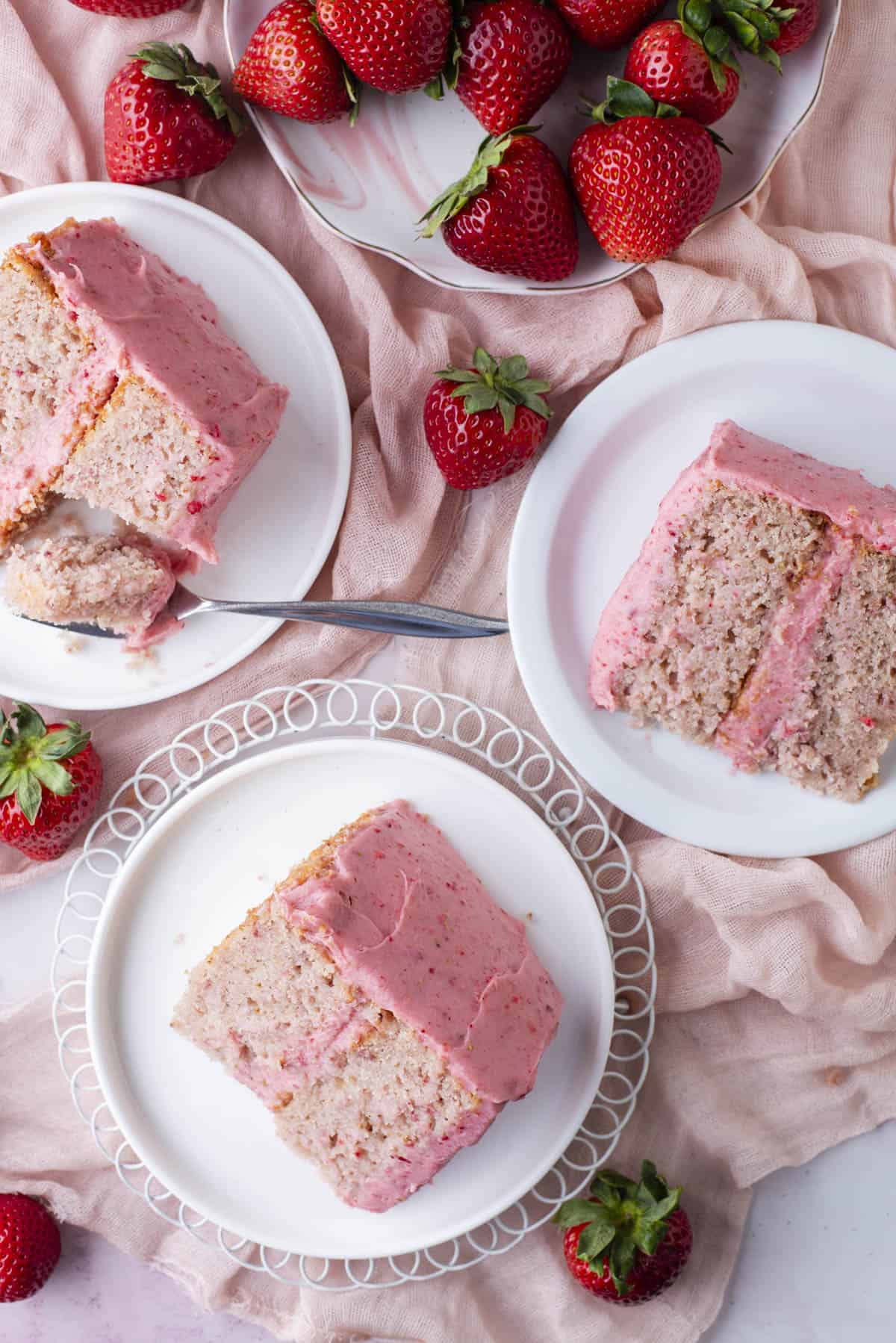 Three plates with slices of strawberry cake topped with pink frosting are arranged on a light pink cloth, surrounded by fresh whole strawberries and a fork cutting into one slice.