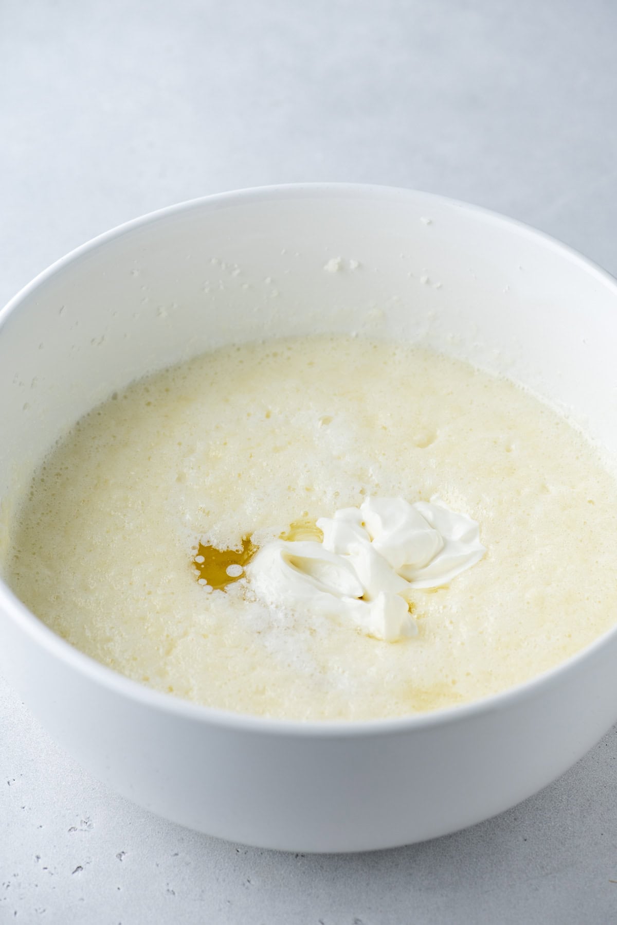 A white mixing bowl filled with a creamy, pale yellow batter, topped with a dollop of sour cream and a small amount of melted butter on a light gray surface.