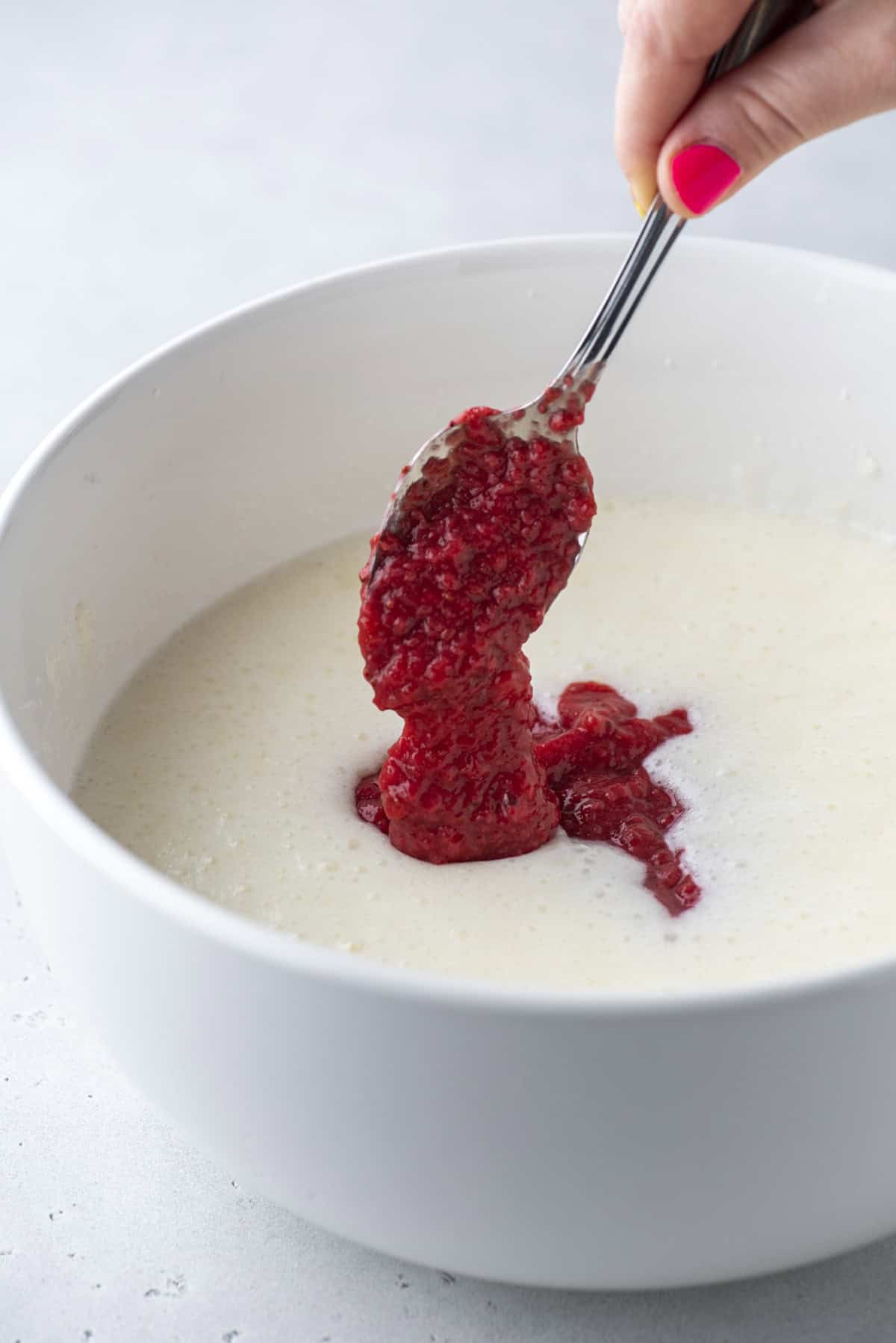 A hand with pink nail polish holds a spoon, adding red raspberry sauce to a white bowl filled with creamy batter.