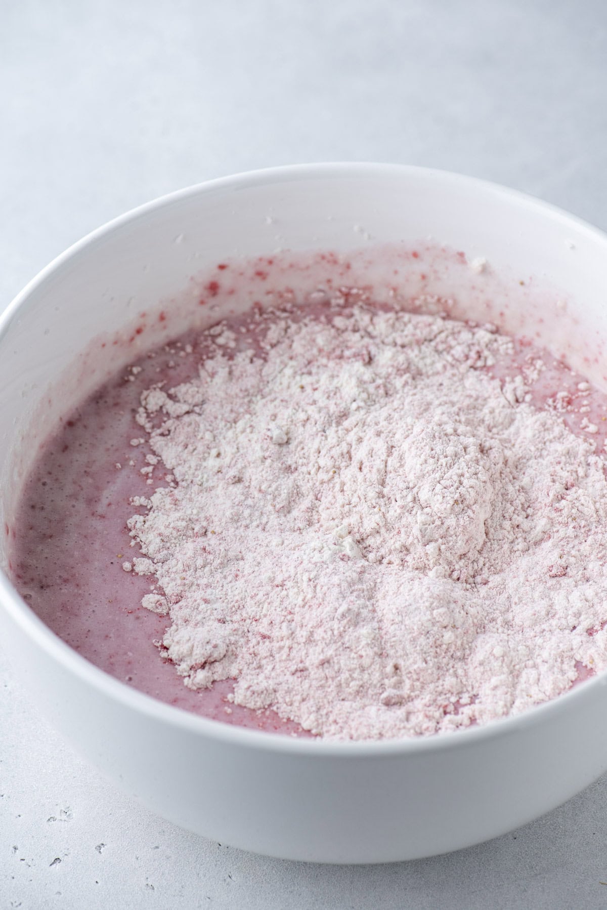 A white mixing bowl filled with pink batter, partially topped with a mound of dry flour mixture, sits on a light gray surface.