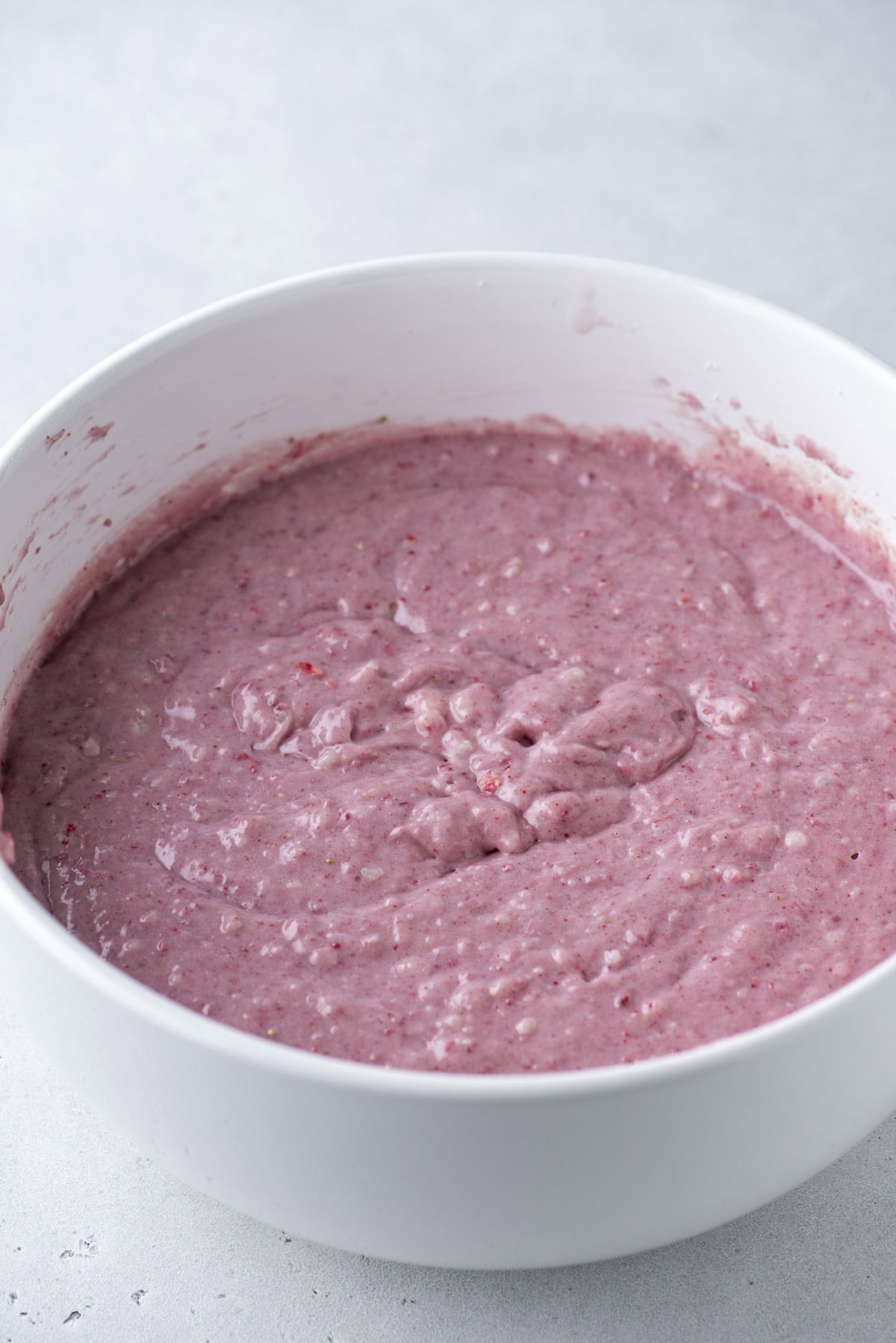 A white mixing bowl filled with thick, pinkish batter, speckled with small bits, sits on a light gray surface.