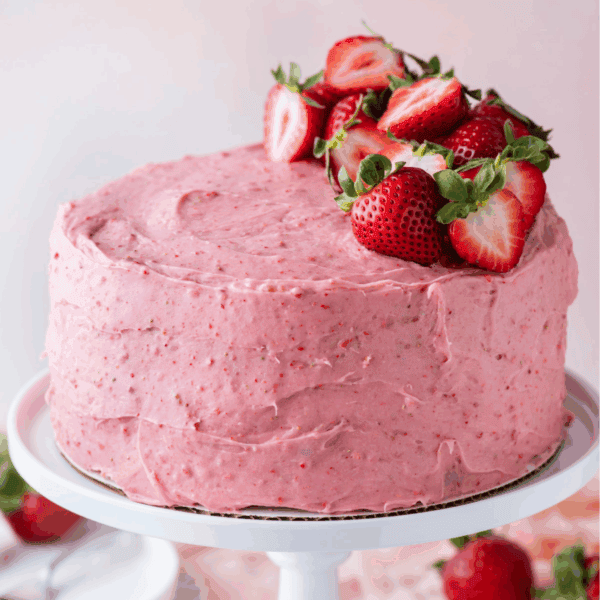 A round cake with pink strawberry frosting sits on a white cake stand, topped with several fresh strawberries, some sliced in half. The background is softly blurred with hints of more strawberries.