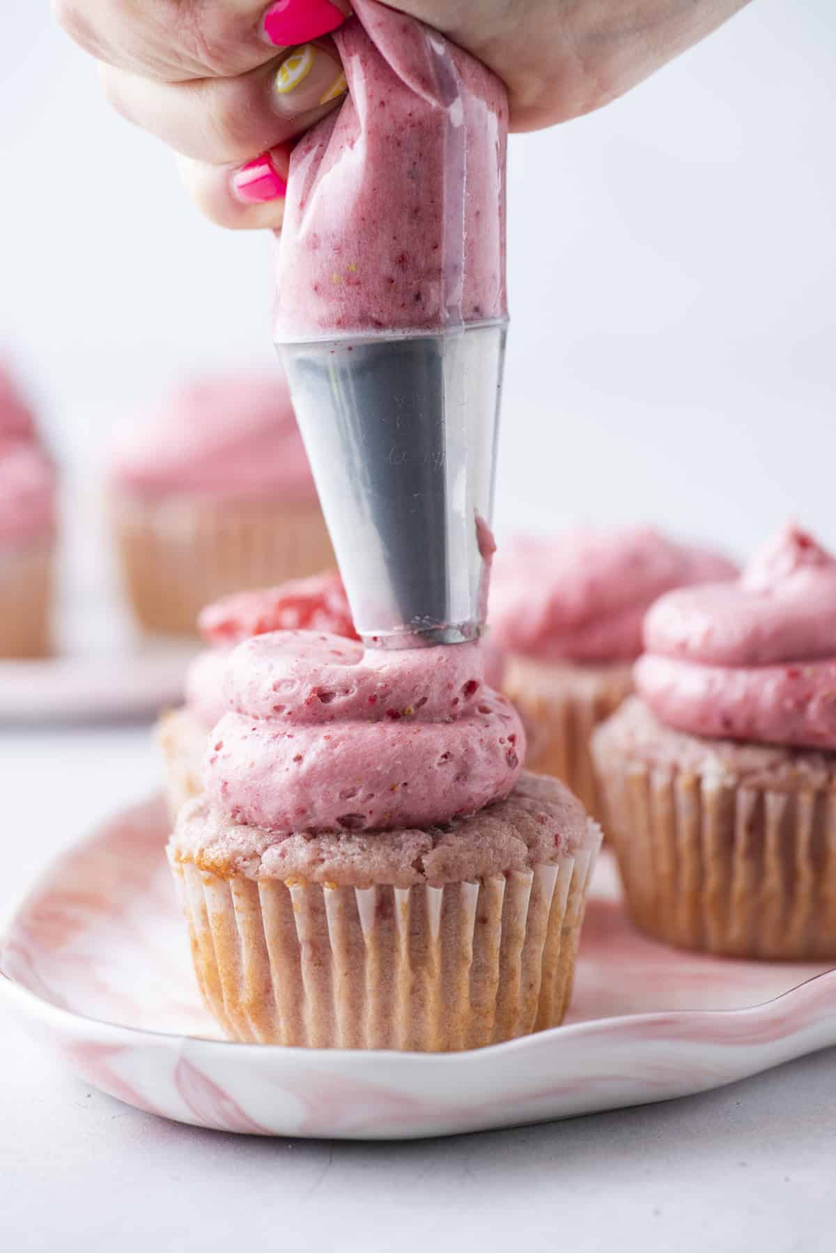 A hand is piping pink strawberry frosting onto a cupcake using a piping bag. Several unfrosted and frosted cupcakes sit on a plate in the background.