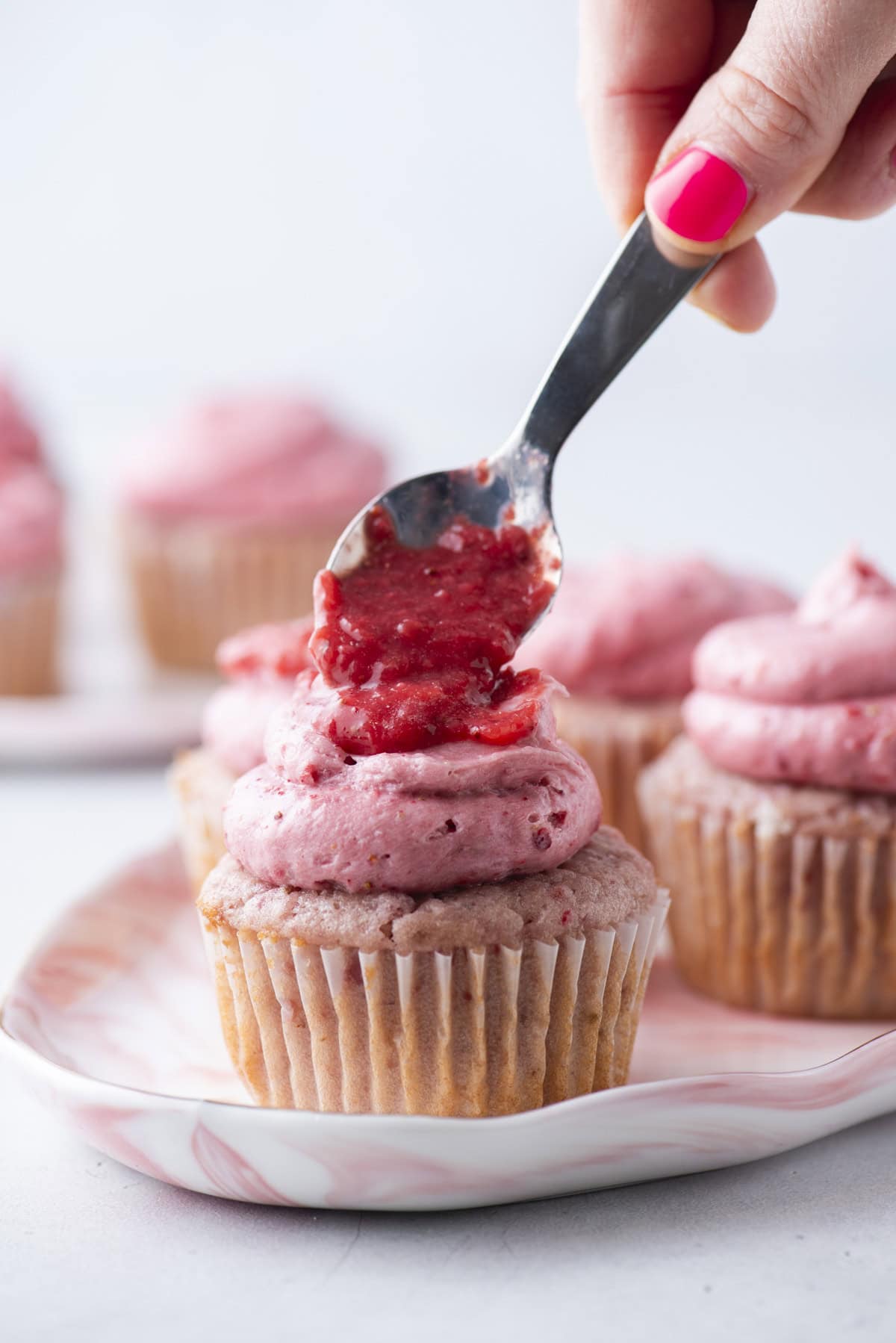 A hand with pink nail polish uses a spoon to spread strawberry topping on a cupcake with pink frosting. More similarly decorated cupcakes are visible in the background on a marbled plate.