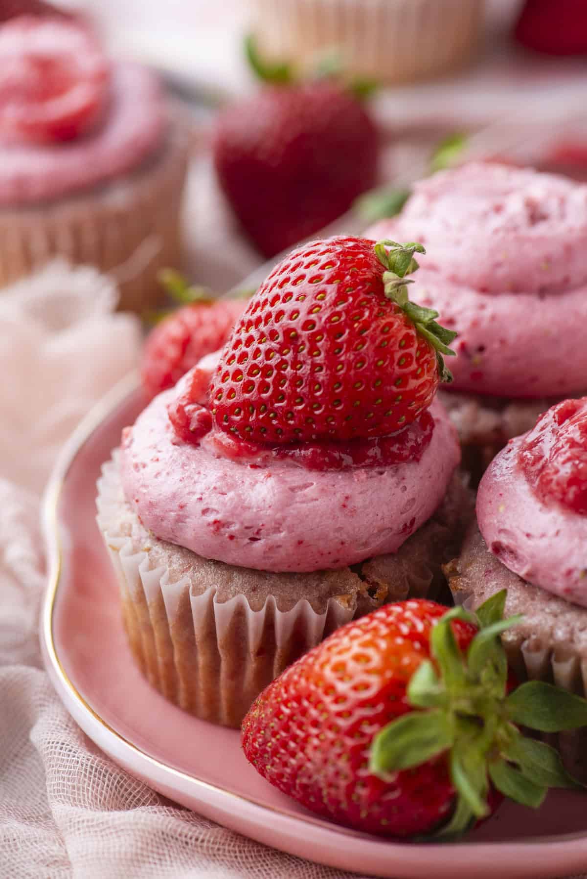Close-up of cupcakes topped with pink strawberry frosting and fresh strawberries, arranged on a pink plate with whole strawberries around them. The background is softly blurred.