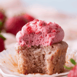 A strawberry cupcake with pink strawberry frosting sits on a white plate. The cupcake has a bite taken out, revealing its moist texture. Fresh strawberries are blurred in the background, and text above reads Strawberry Cupcakes.