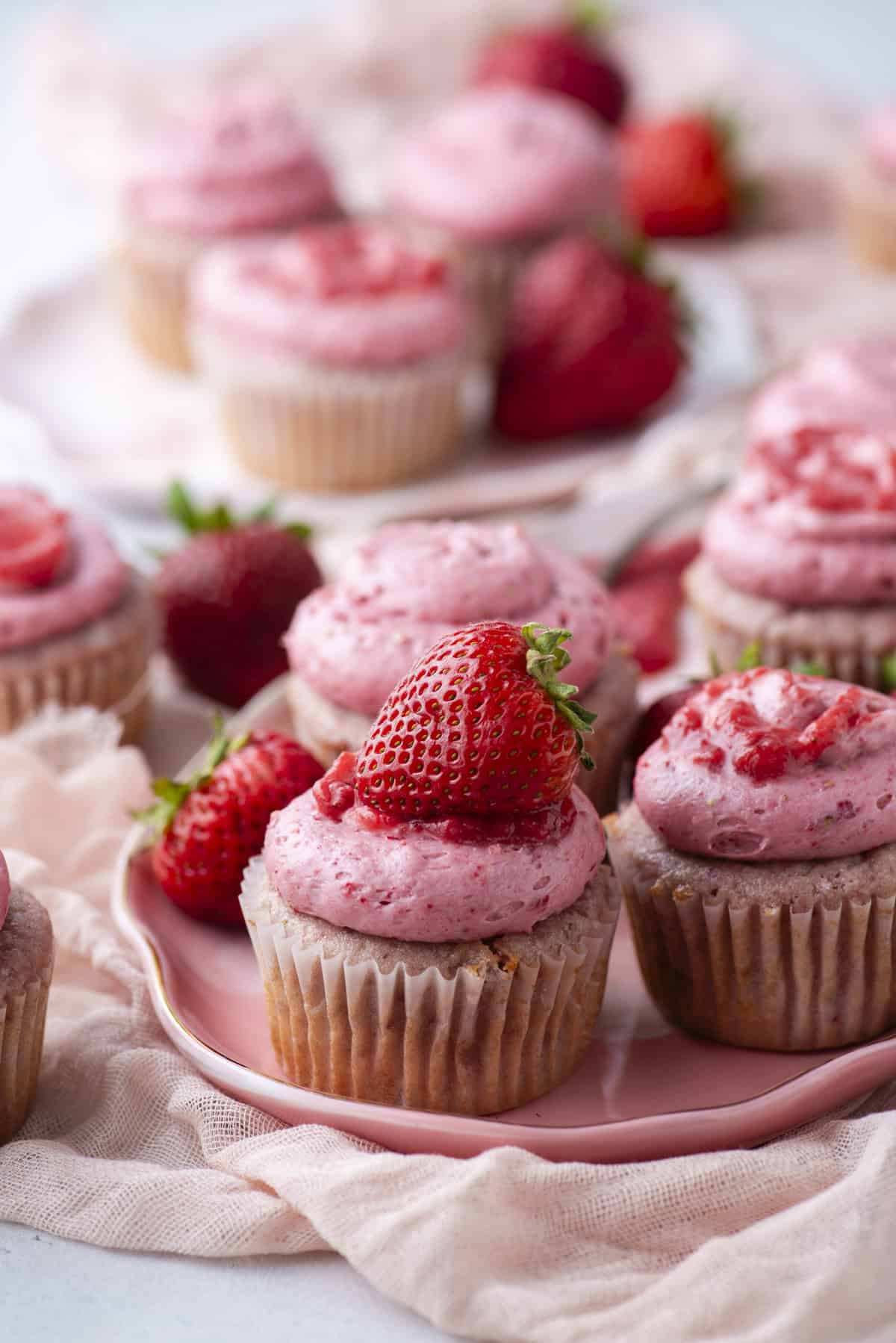 Cupcakes topped with pink strawberry frosting and fresh strawberries are arranged on a pink plate, with more cupcakes and whole strawberries in the background. A soft, light fabric is draped around the plate.