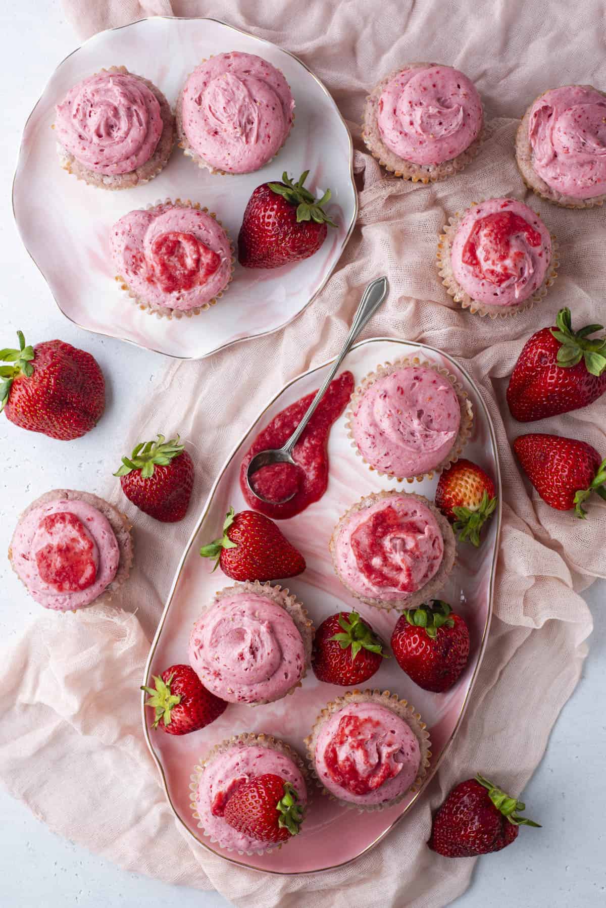 An overhead view of strawberry cupcakes with pink frosting on decorative plates, surrounded by fresh strawberries and a pink cloth, with a spoon holding strawberry sauce.