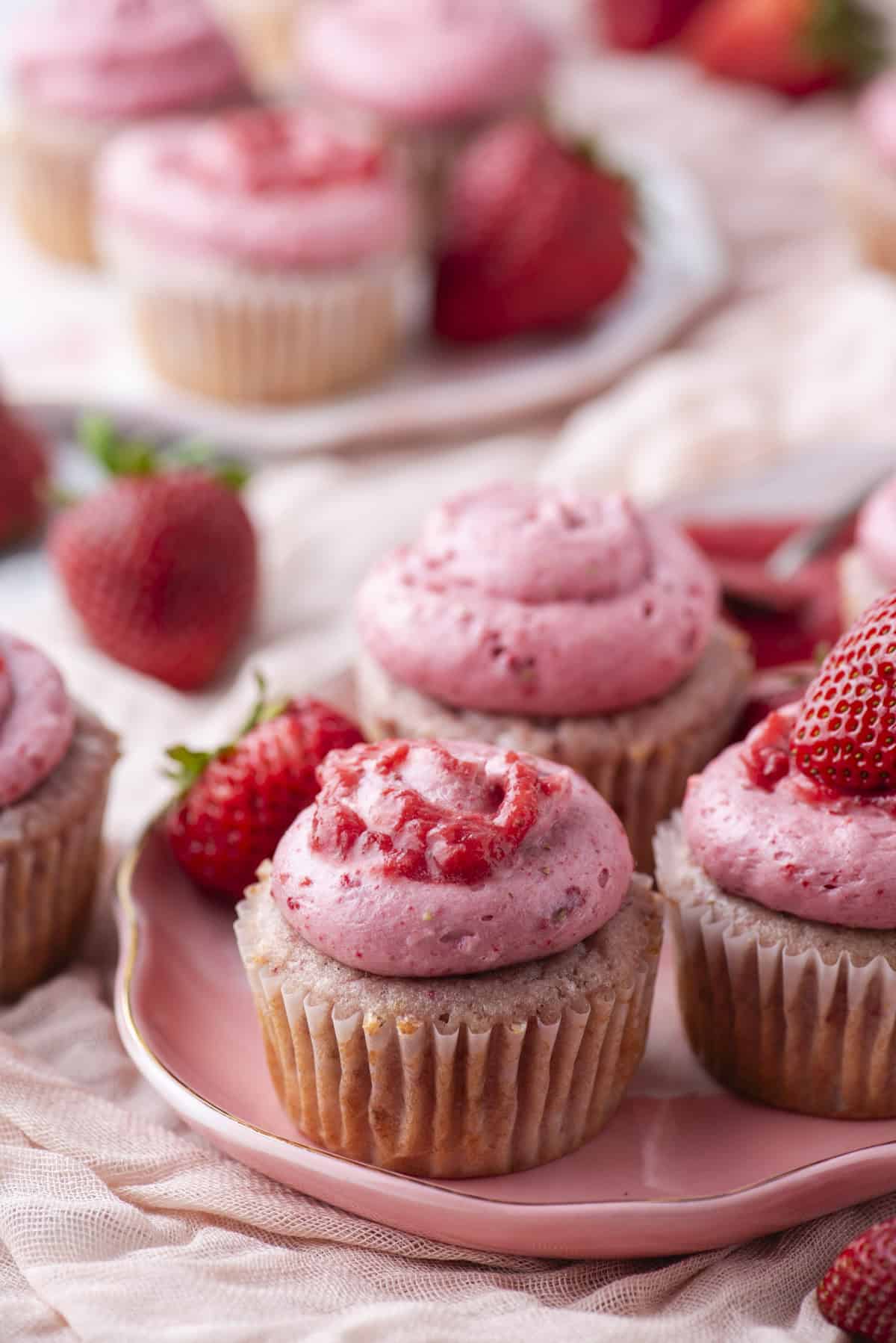 A close-up of vanilla cupcakes topped with pink strawberry frosting and fresh strawberries, arranged on a pink plate with whole strawberries scattered around.