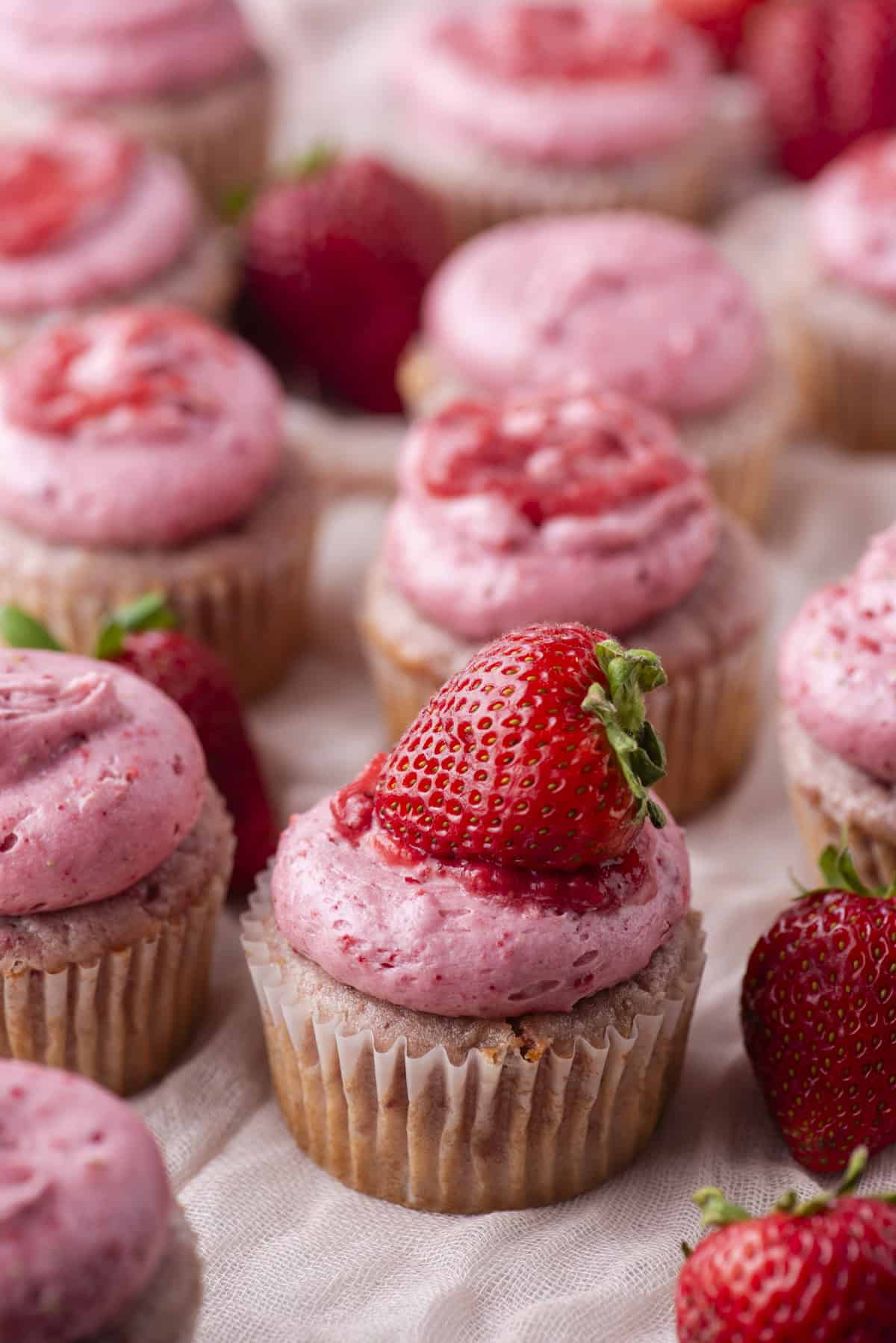 Cupcakes topped with pink strawberry frosting are arranged in rows. Some cupcakes are decorated with fresh strawberries and a drizzle of strawberry sauce. Whole strawberries are scattered among the cupcakes.