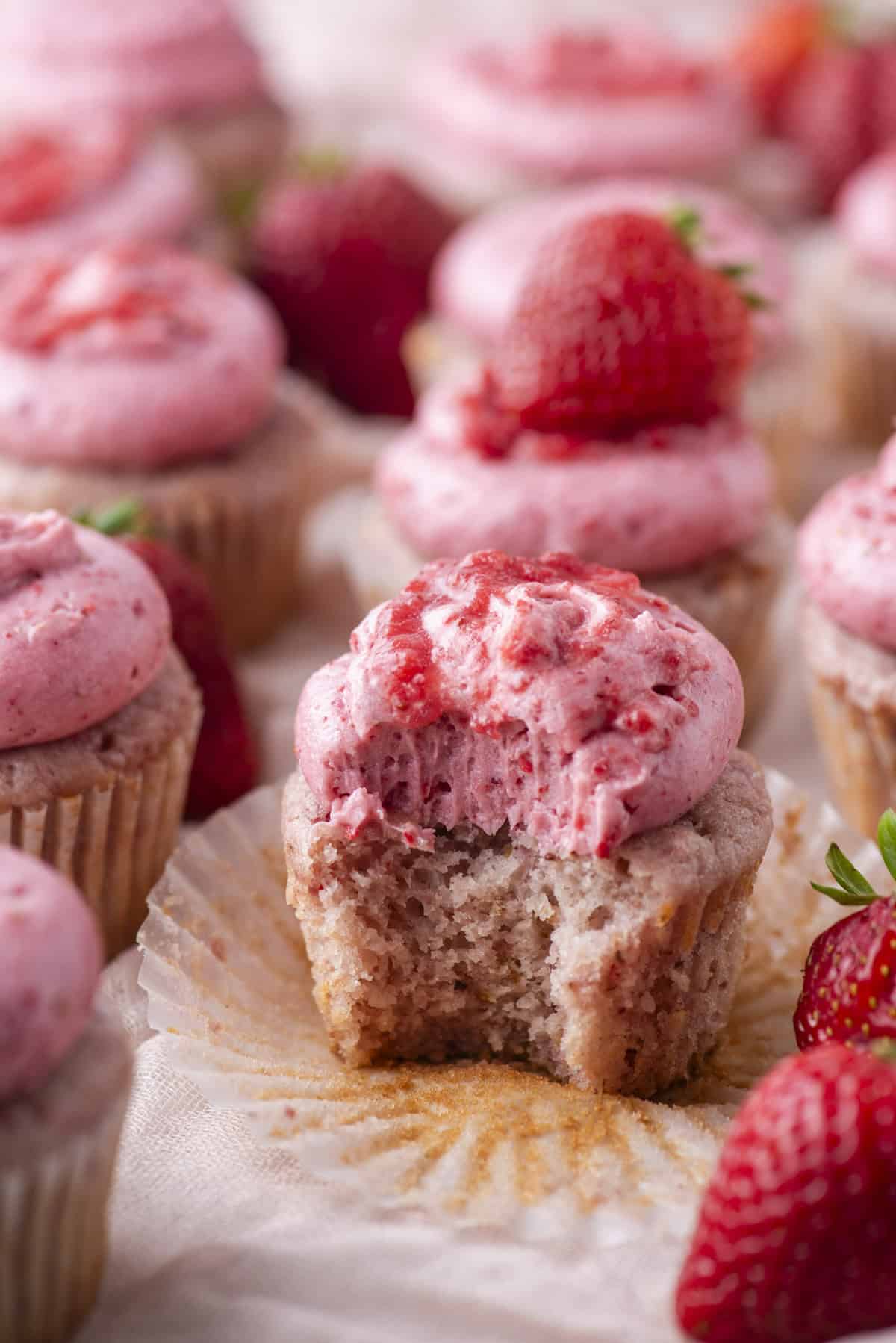 A close-up of strawberry cupcakes with pink frosting and fresh strawberries on top. One cupcake in the center has a bite taken out, revealing its moist interior. Cupcakes and strawberries are scattered in the background.