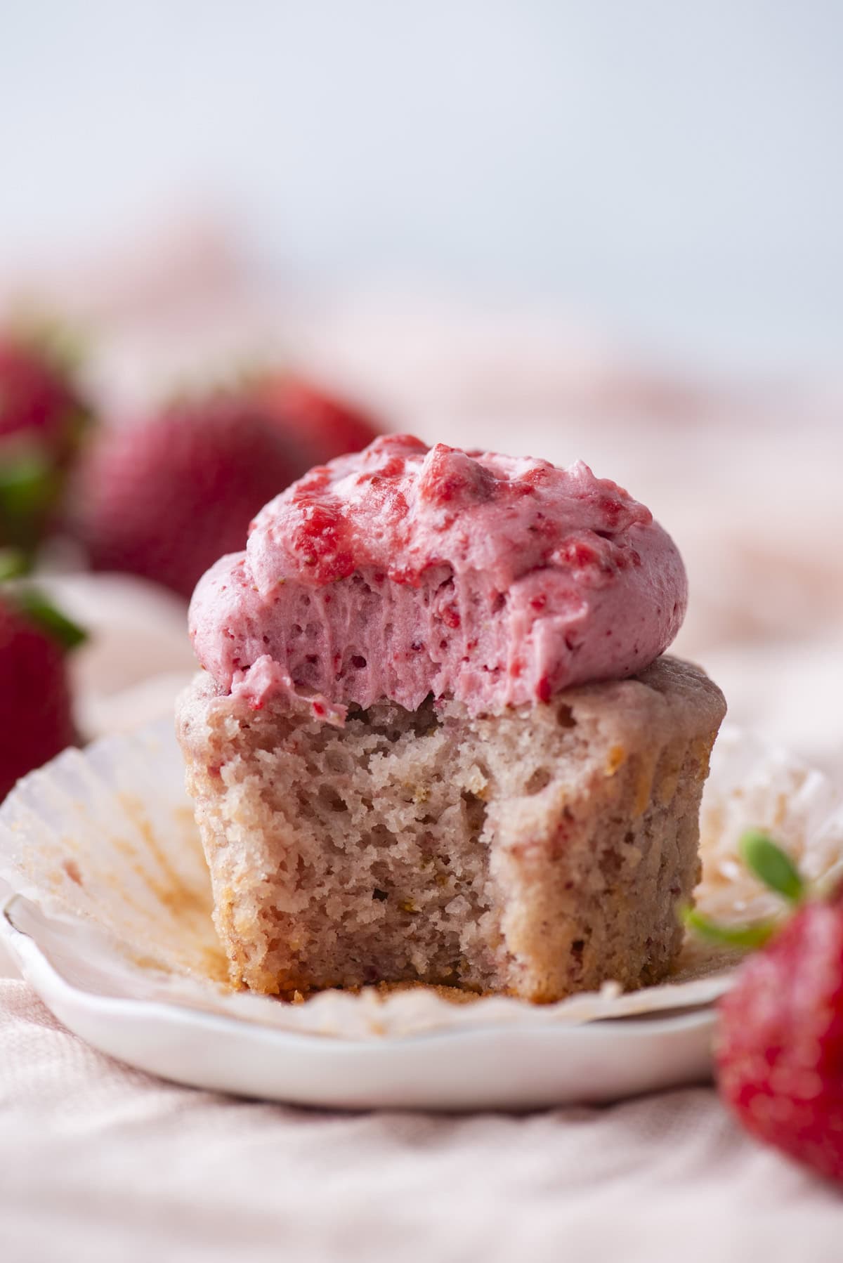 A strawberry cupcake with pink strawberry frosting, partially unwrapped and with a bite taken out, sits on a small plate. Fresh strawberries are blurred in the background.