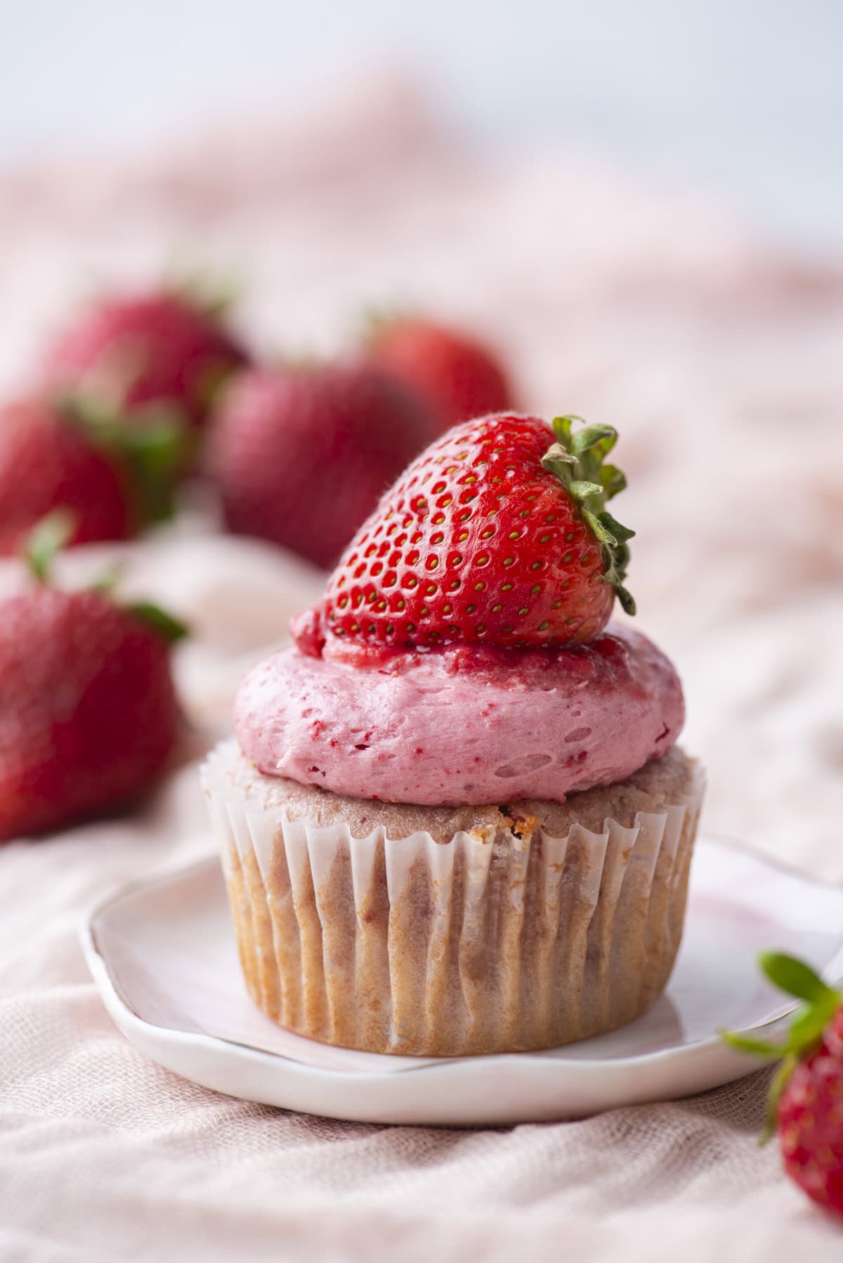 A cupcake with pink strawberry frosting, topped with a fresh whole strawberry, sits on a small white plate. Blurred strawberries are visible in the background.