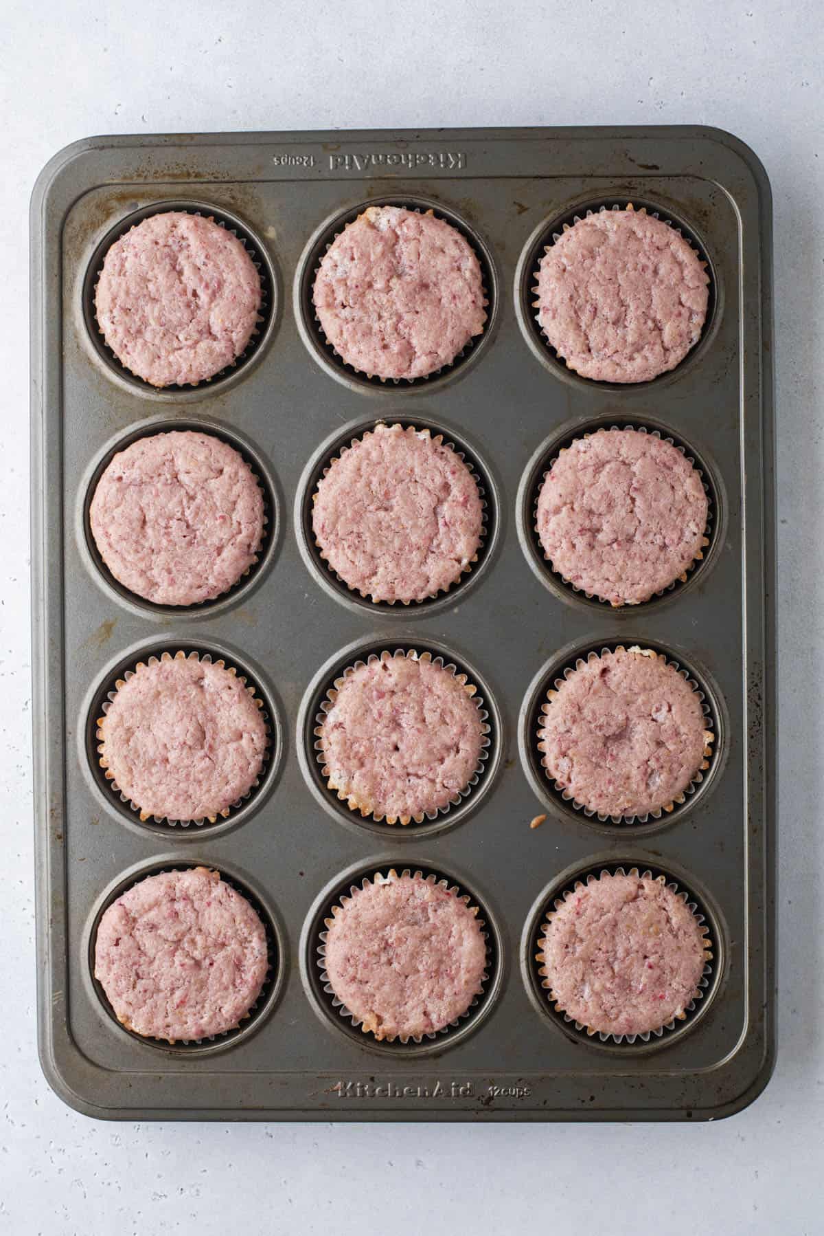 A muffin tin with twelve cupcake liners filled with baked, pinkish muffins, viewed from above on a light gray surface.