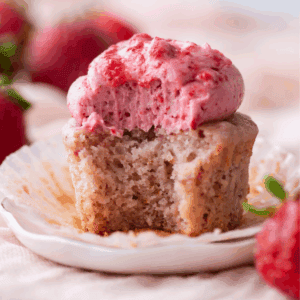 A partially eaten strawberry cupcake with pink strawberry frosting sits on a small white plate, surrounded by fresh strawberries. The cupcake’s liner is peeled back, revealing its moist, textured interior.