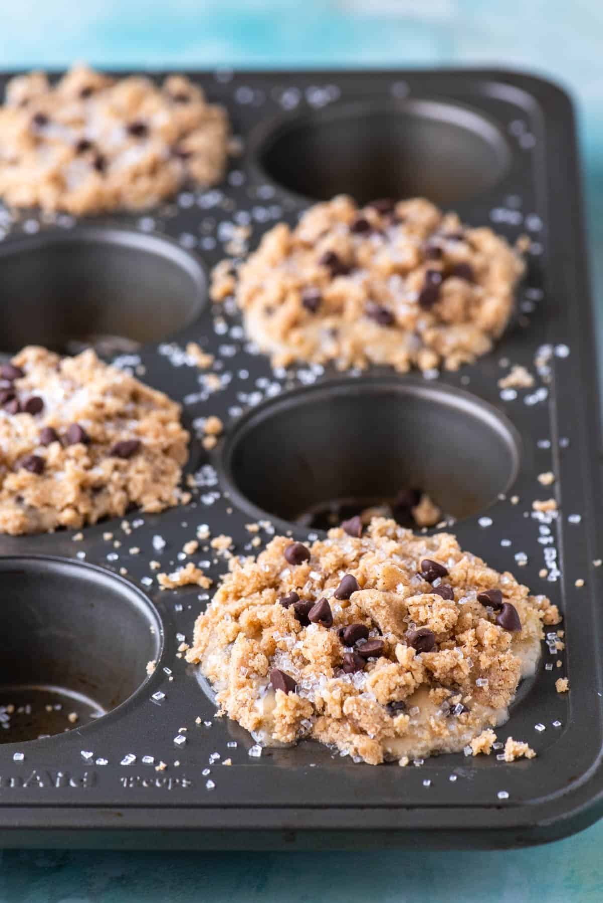 A muffin tin filled with unbaked muffins topped with crumbly streusel and chocolate chips, with some salt sprinkled on top. The tin has empty spaces and sits on a light blue surface.