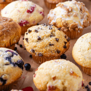A variety of muffins, including ones with chocolate chips, berries, cinnamon sugar, and crumb toppings, arranged closely together on a brown surface.