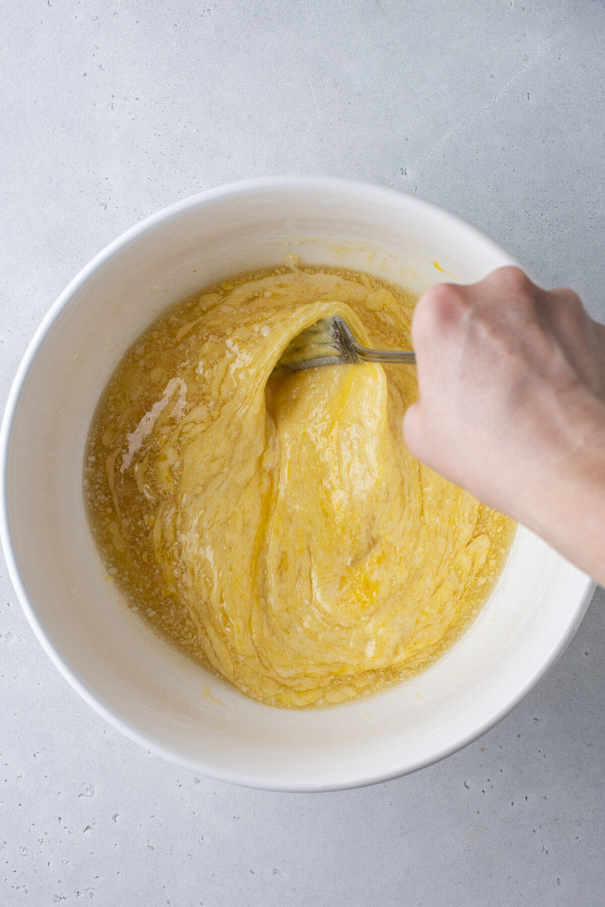 A hand uses a fork to mix yellow batter with specks of black seasoning in a white bowl on a light gray surface.