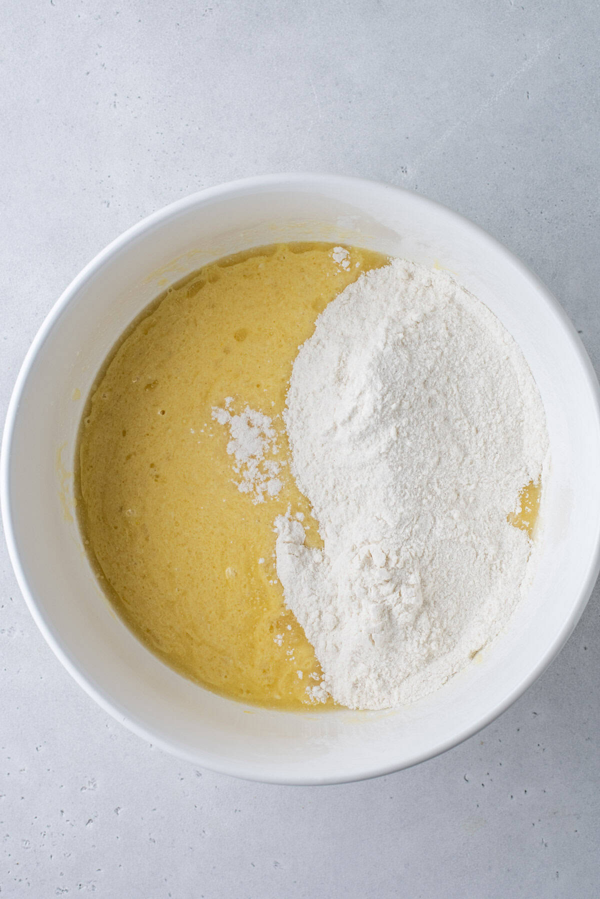 A white bowl containing a yellow liquid batter and a pile of white flour, partially mixed together, sits on a light gray surface.