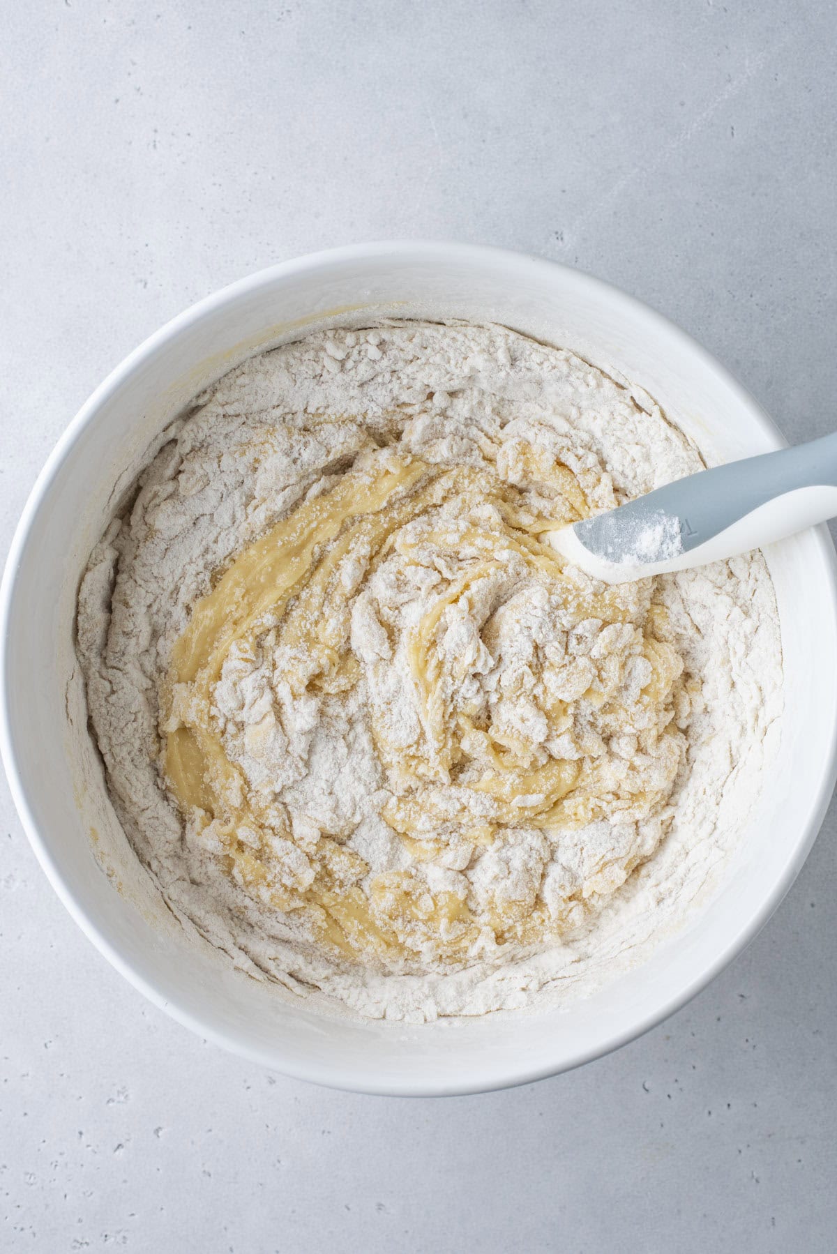 A white bowl containing partially mixed dough with visible dry flour and wet ingredients, and a gray spatula resting on the side of the bowl, on a light gray surface.