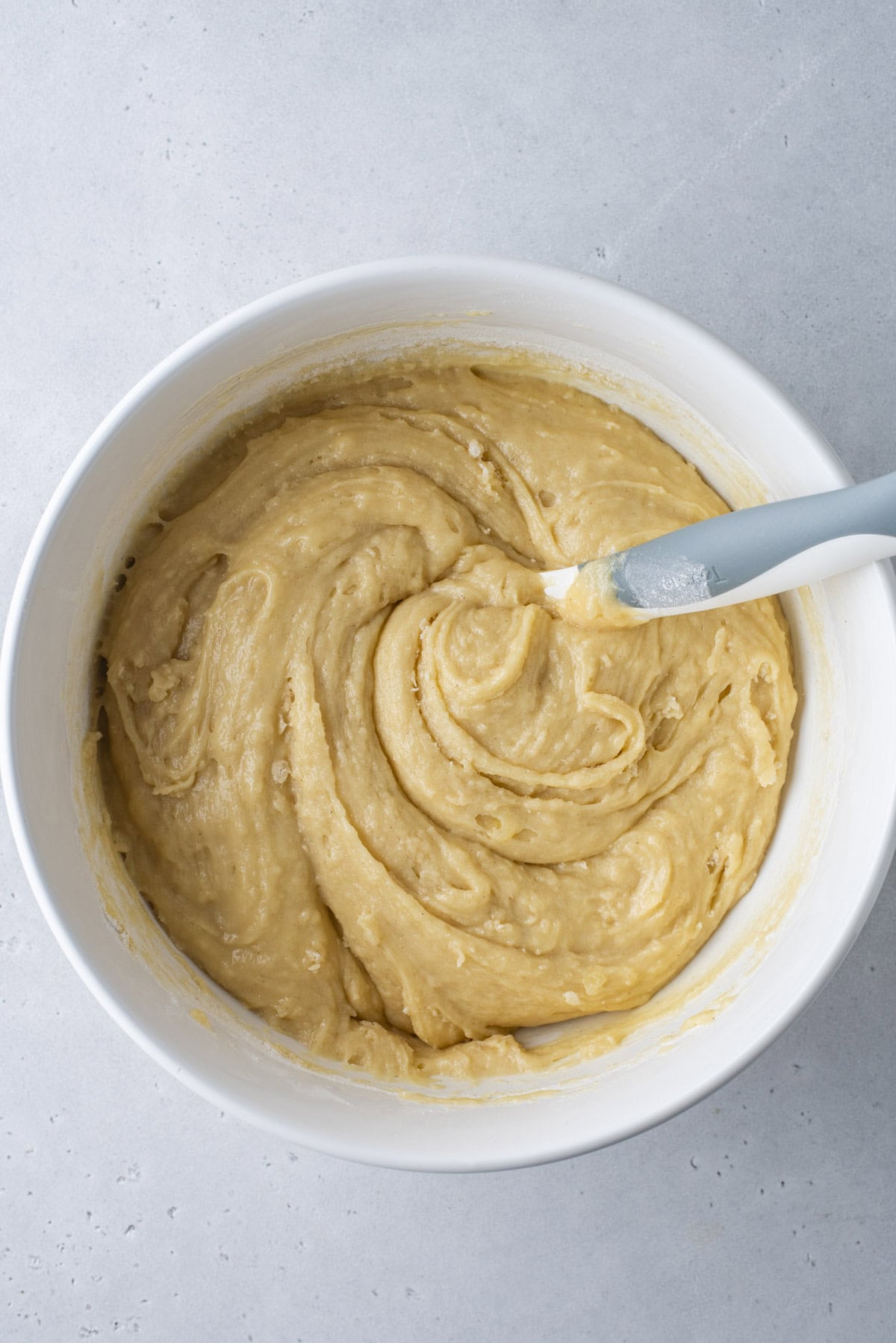 A white bowl filled with creamy, light yellow batter being mixed with a blue and white spatula on a light gray surface.