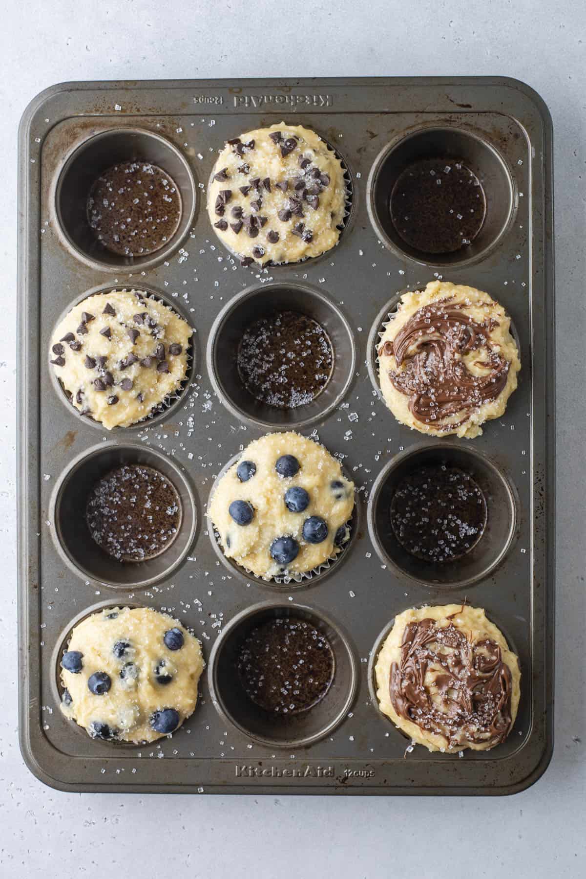A muffin tin holds six unbaked muffins topped with chocolate chips, blueberries, or chocolate swirls, while three cupcake slots are empty, showing only the liners and sprinkled salt.