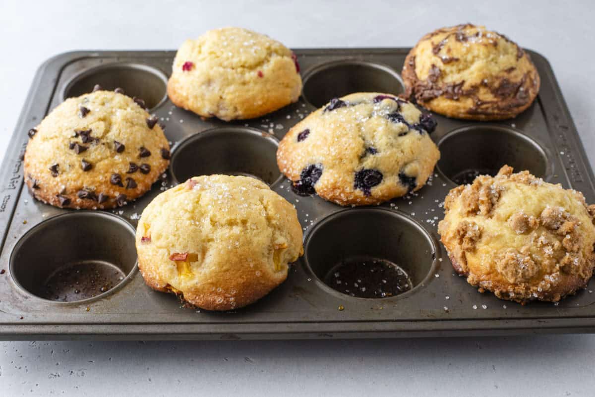 A metal muffin tray holding six assorted muffins, including ones with chocolate chips, blueberries, and crumble toppings, on a light gray surface.
