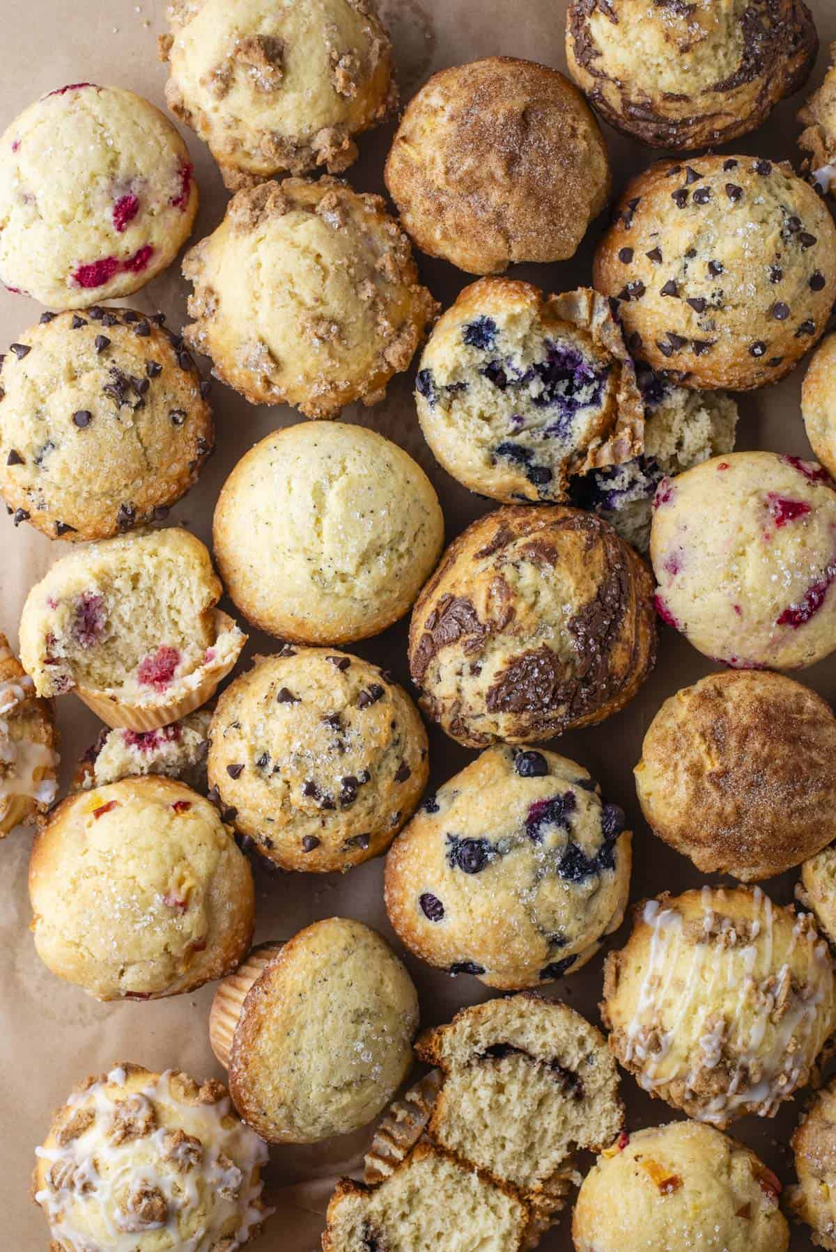 An assortment of muffins, including blueberry, chocolate chip, and streusel-topped varieties, arranged closely together on a parchment surface, some whole and some cut open to reveal their fillings.