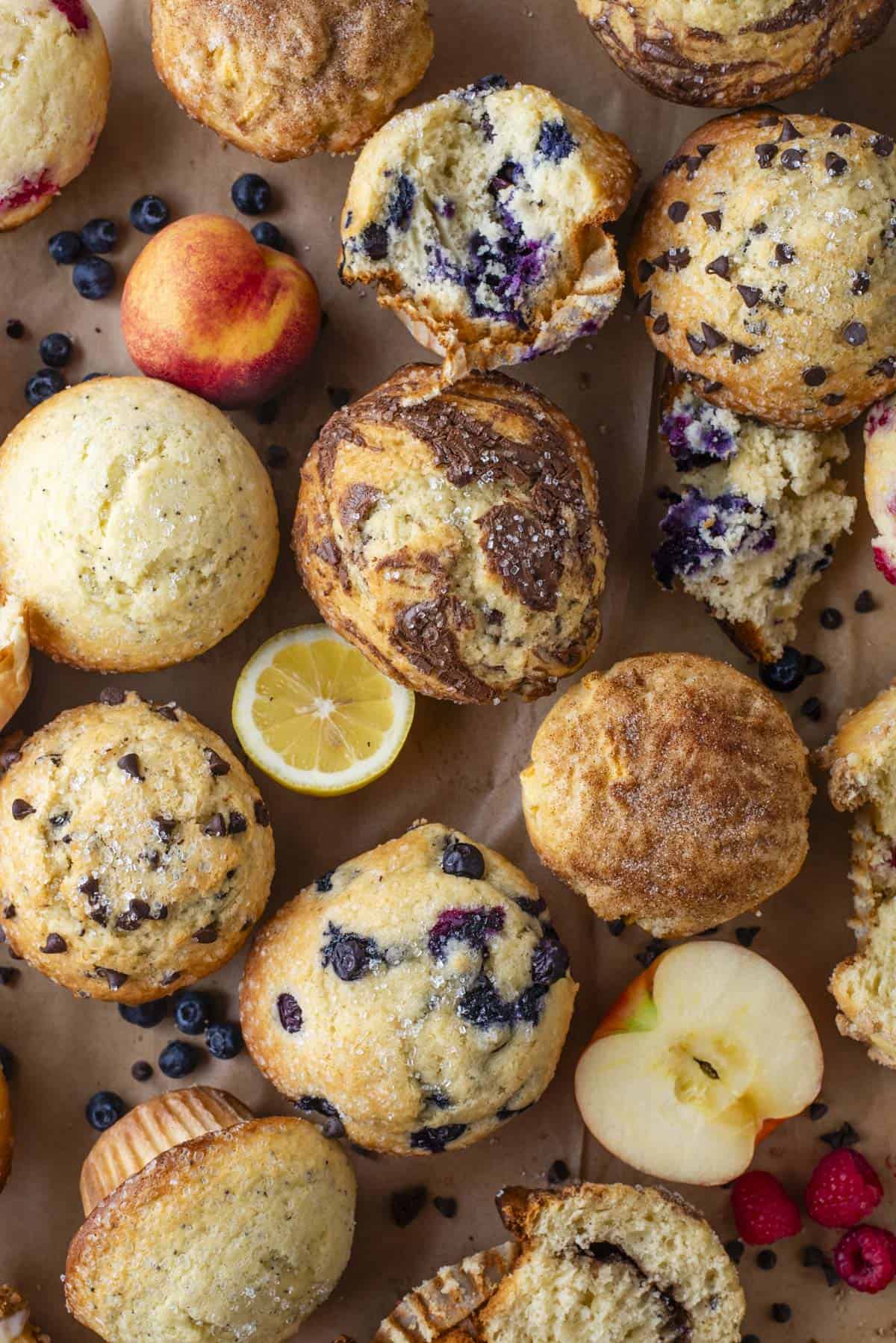 An assortment of muffins with various mix-ins like blueberries and chocolate chips, surrounded by fresh fruit including peaches, a lemon slice, apples, and scattered blueberries on brown parchment paper.