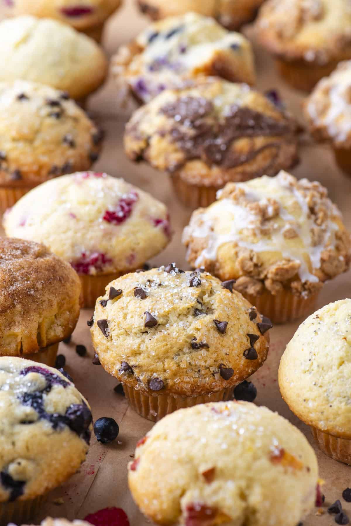 An assortment of freshly baked muffins, including chocolate chip, blueberry, and streusel-topped varieties, arranged closely on a brown parchment surface.