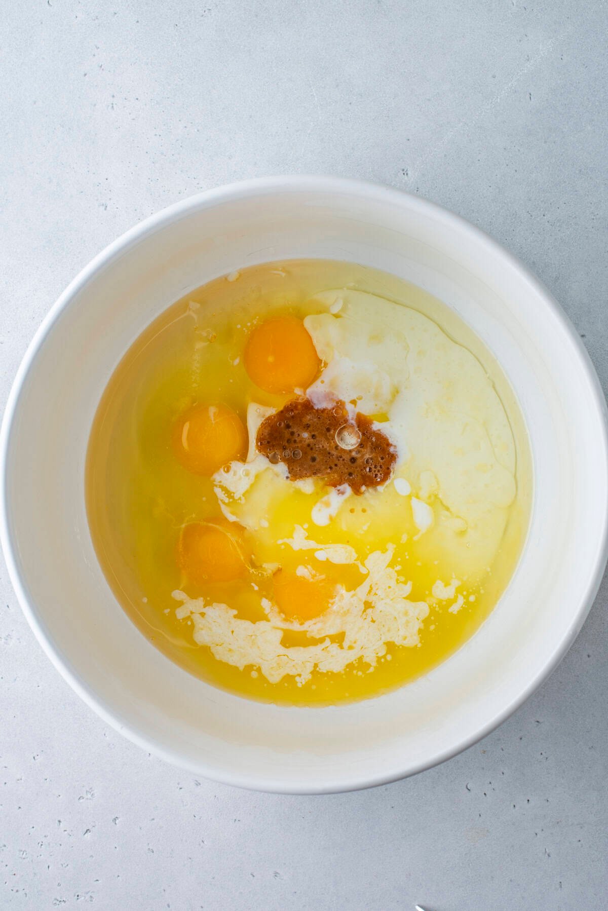 A white mixing bowl on a gray surface containing oil, vinegar, spices, and seasonings before being mixed together.