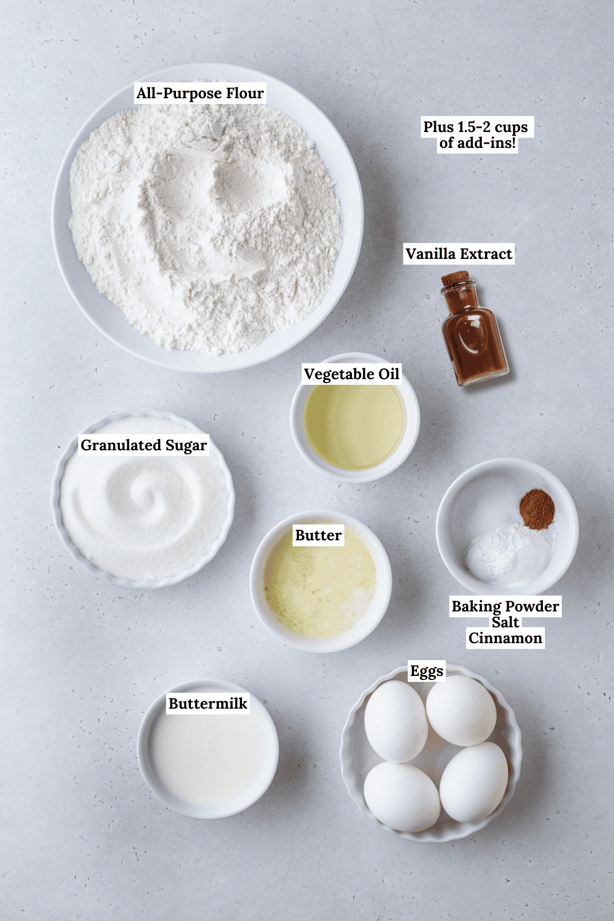 Top-down view of labeled baking ingredients in bowls: all-purpose flour, granulated sugar, buttermilk, eggs, melted butter, vegetable oil, vanilla extract, and a small bowl with baking powder, salt, and cinnamon.