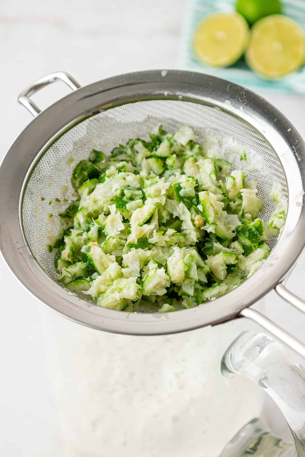 A metal strainer filled with finely chopped green and white vegetables sits atop a glass pitcher. In the background, there are halved limes on a plate.