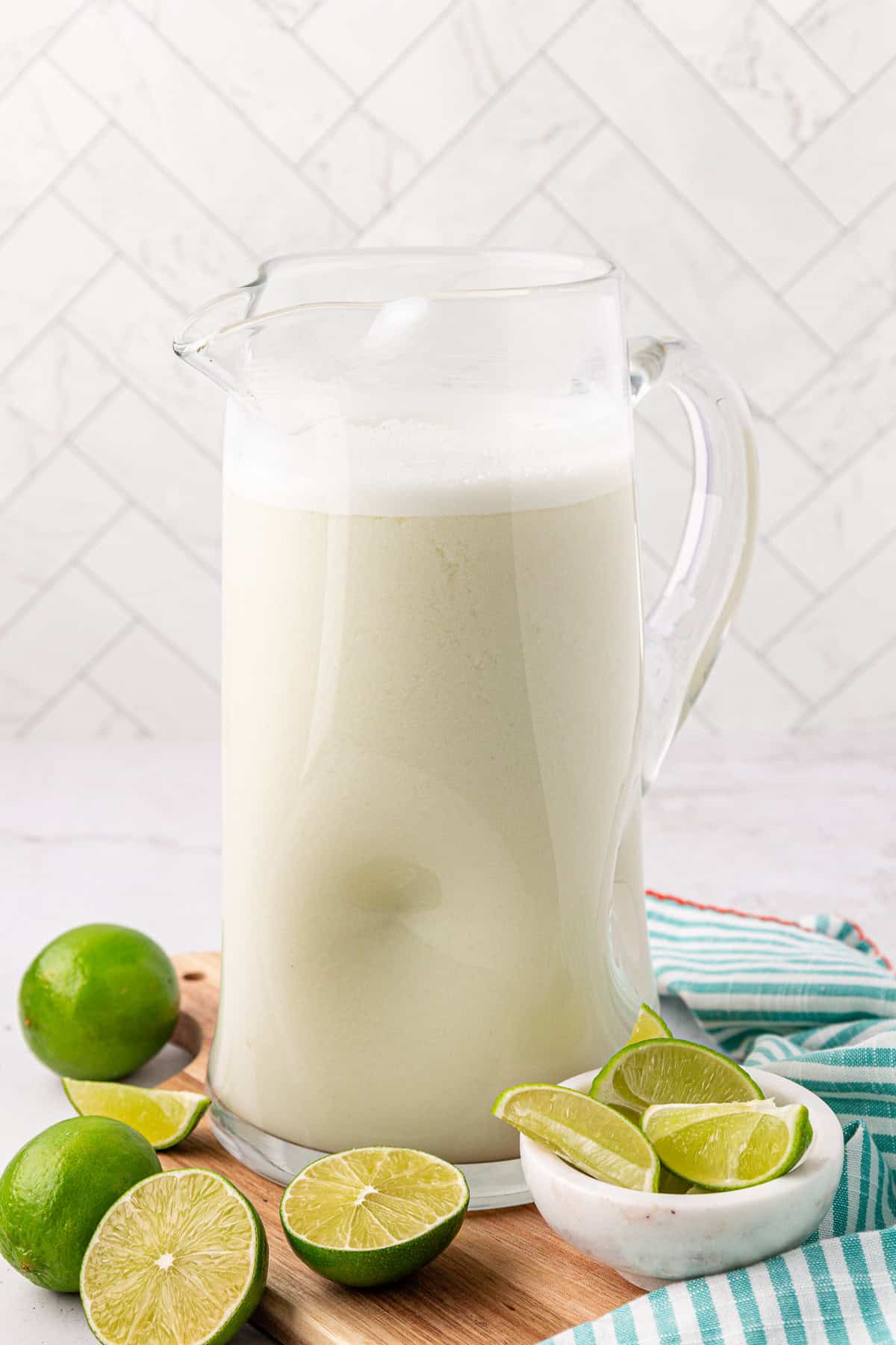 A glass pitcher filled with a frothy, creamy white beverage sits on a wooden board, surrounded by whole and halved limes. A striped cloth is nearby, and the background features white tiled walls.