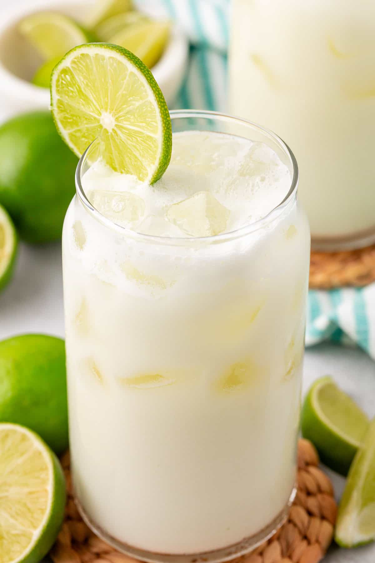 A glass of iced, creamy white lime drink garnished with a slice of lime sits on a woven coaster. Fresh limes and lime wedges surround the glass, and a bowl of limes is in the background.