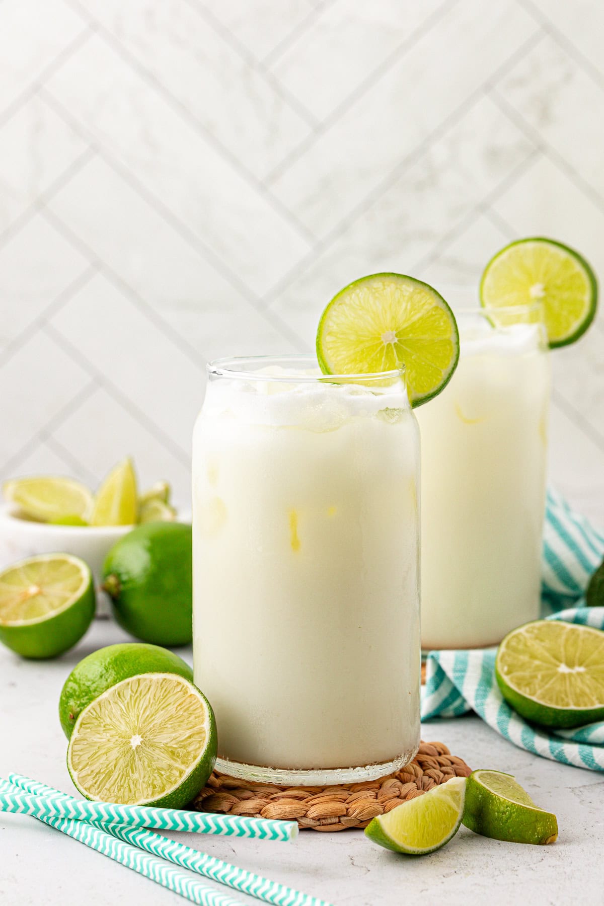 Two glasses of a creamy, iced white drink garnished with lime slices, surrounded by fresh cut limes and blue striped straws on a light surface with a white tiled background.