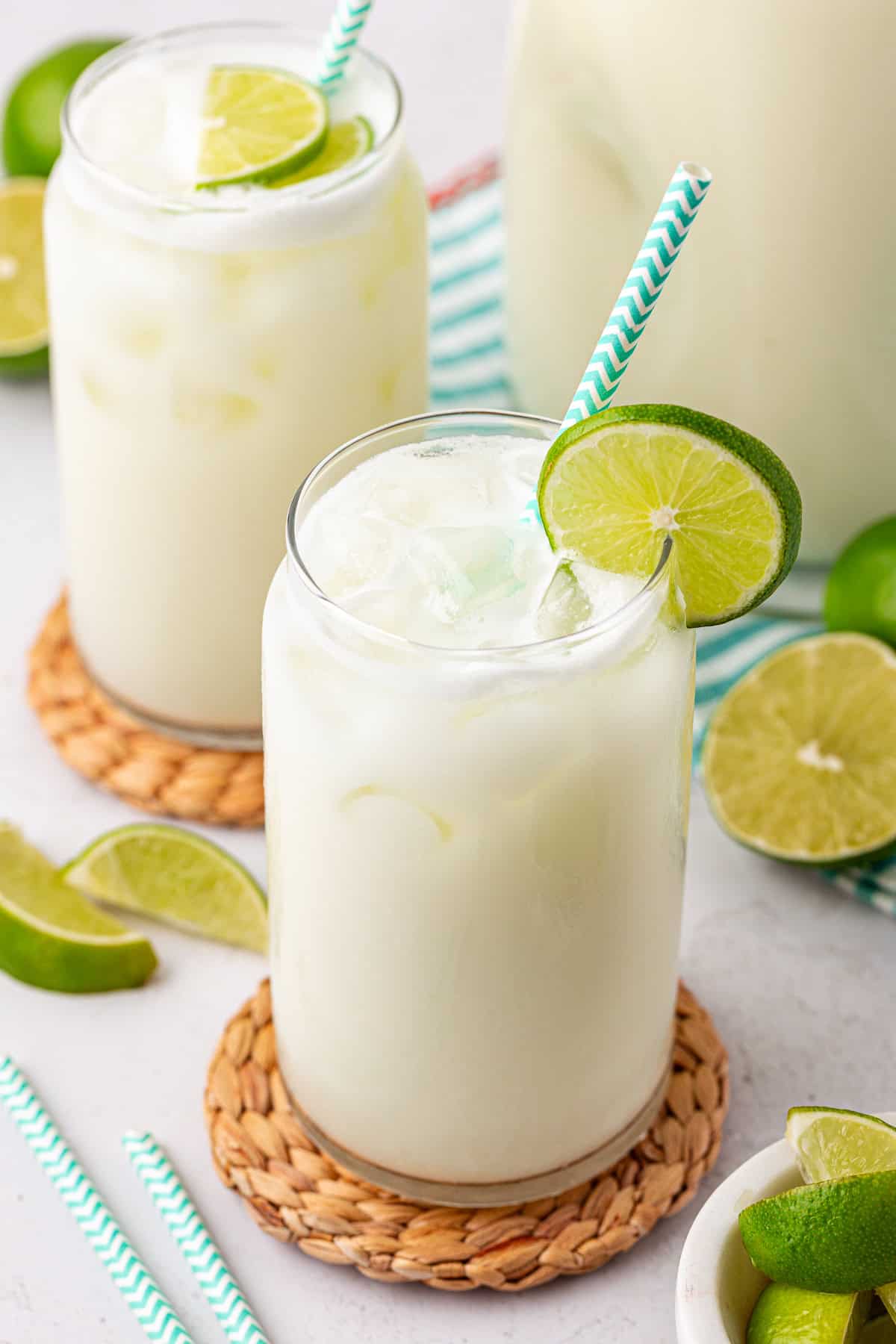 Two glasses of creamy, iced limeade with green-and-white striped straws, garnished with lime slices, sit on woven coasters. Fresh lime wedges are scattered around on a light surface.