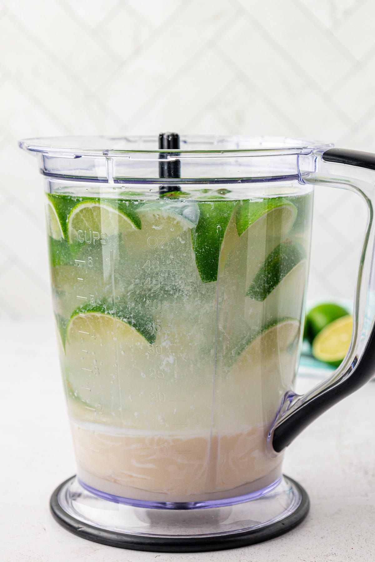 A blender pitcher filled with water, lime wedges, and a cloudy liquid, sitting on a white surface with a white tiled background.
