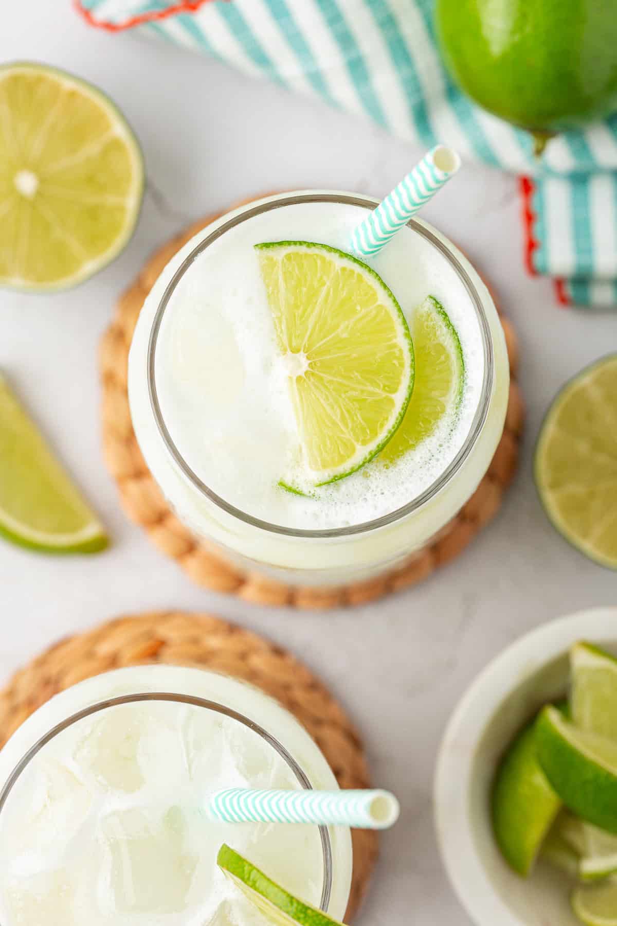 Two glasses filled with ice and a light, frothy drink, each garnished with lime slices and a striped straw, surrounded by additional lime wedges on a white surface.