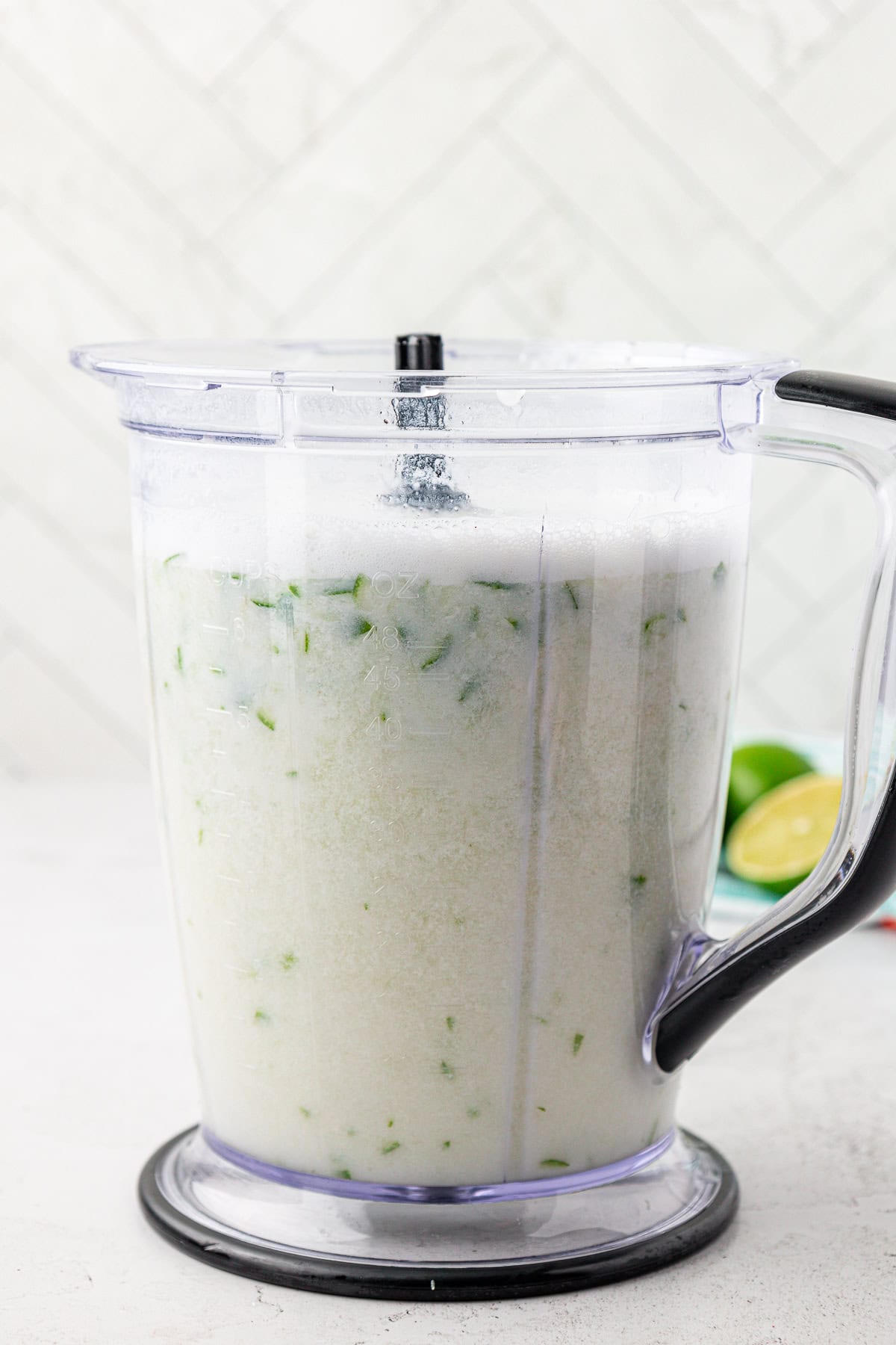 A blender filled with a frothy, light-colored liquid containing small green pieces, sitting on a white surface. Lime halves are visible in the background.