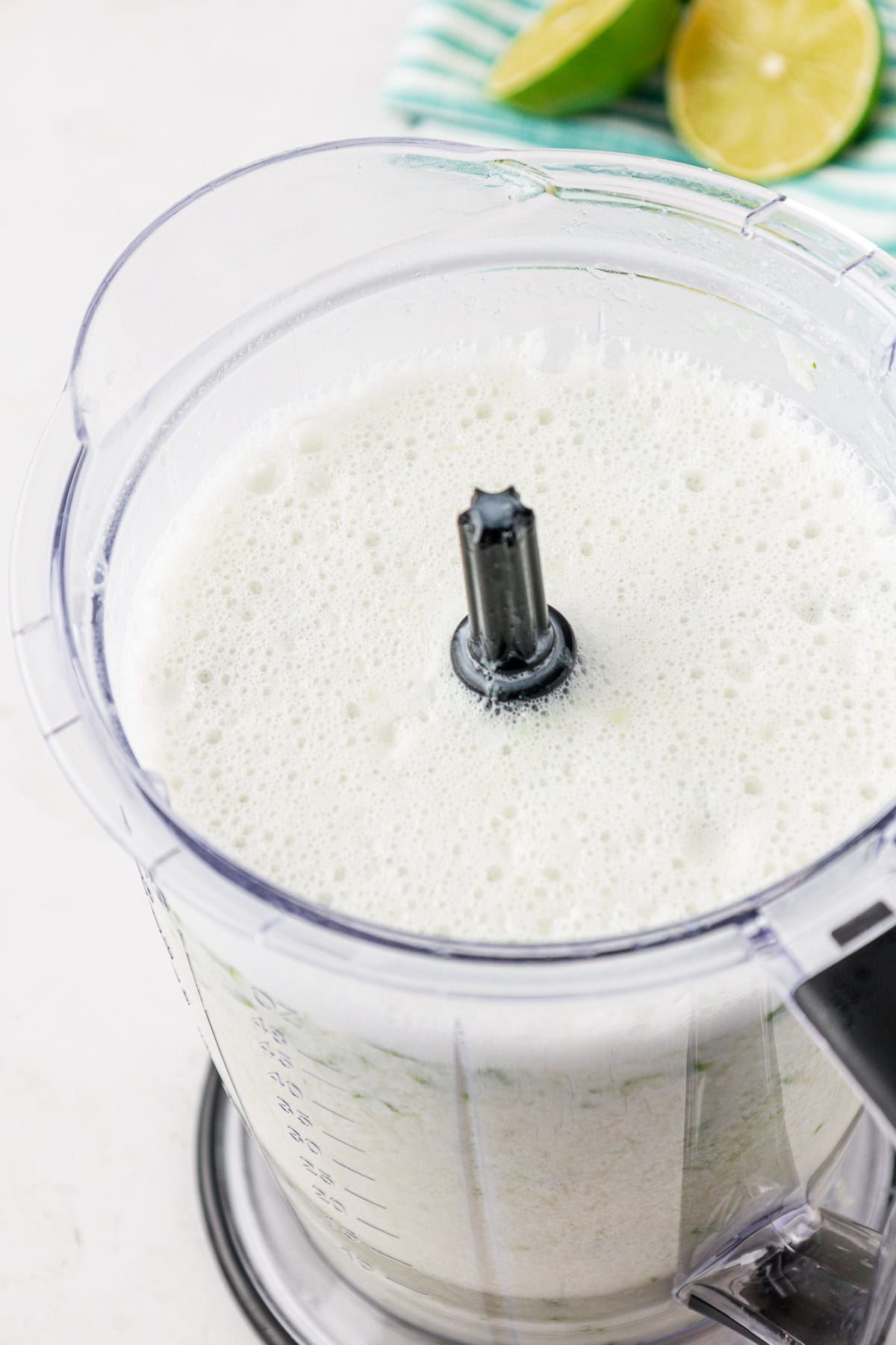 A blender filled with a frothy white liquid, with fresh lime halves and a striped towel in the background.