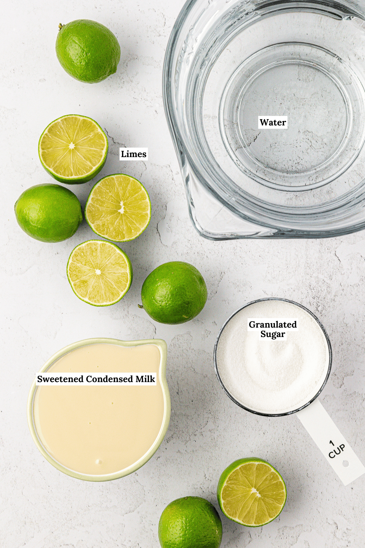 Top-down view of ingredients for brazilian lemonade: a bowl of water, several whole and halved limes, a measuring cup of granulated sugar, and a bowl of sweetened condensed milk on a white surface. Labels identify each item.