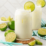 Two glasses of a creamy white drink with ice, each garnished with a lime slice, sit on a woven mat. Fresh limes and blue-striped straws are nearby, with a white tiled background.