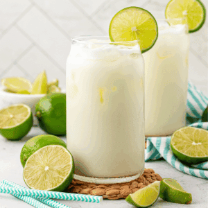 Two glasses of a creamy white drink with ice, each garnished with a lime slice, sit on a woven mat. Fresh limes and blue-striped straws are nearby, with a white tiled background.