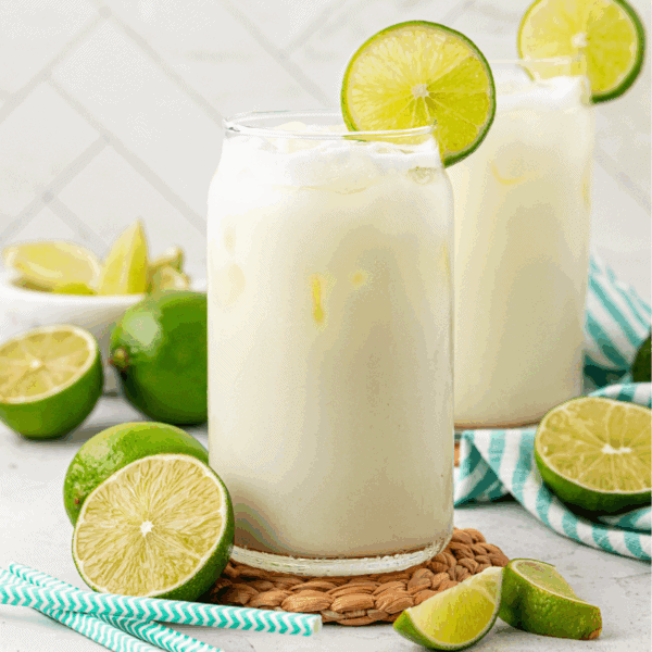Two glasses of a creamy white drink with ice, each garnished with a lime slice, sit on a woven mat. Fresh limes and blue-striped straws are nearby, with a white tiled background.